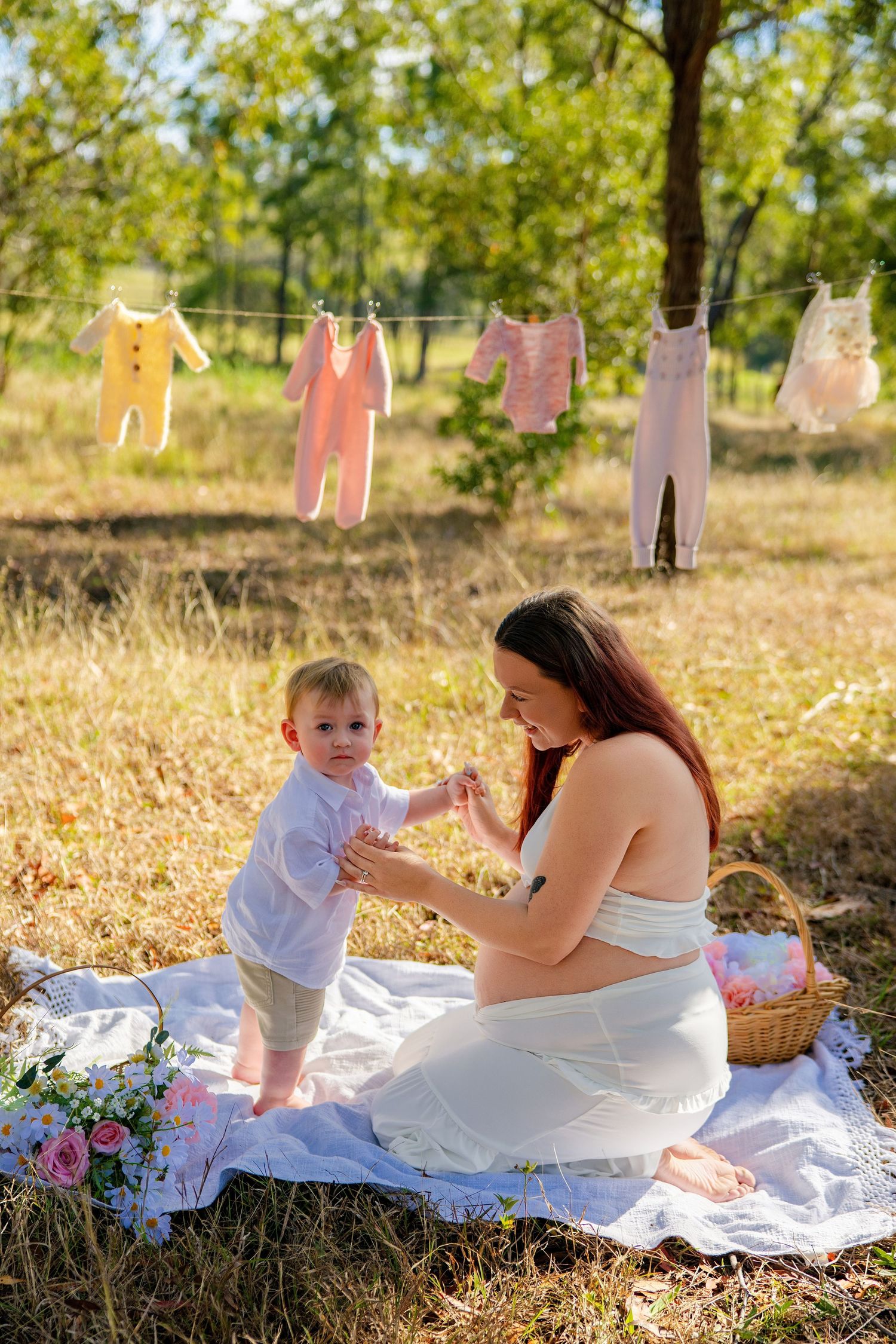 Pregnant woman in white dress plays with toddler on blanket outdoors, baby clothes hanging on line in sunny garden.