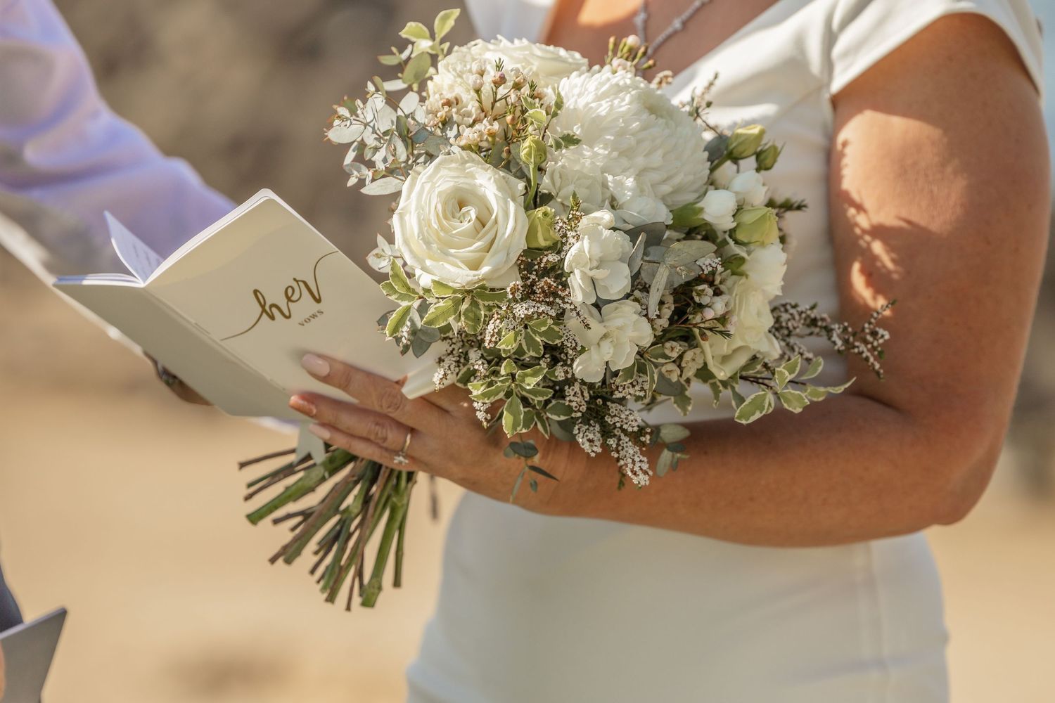 Close-up view of hands holding a white rose and eucalyptus wedding bouquet against a light background.