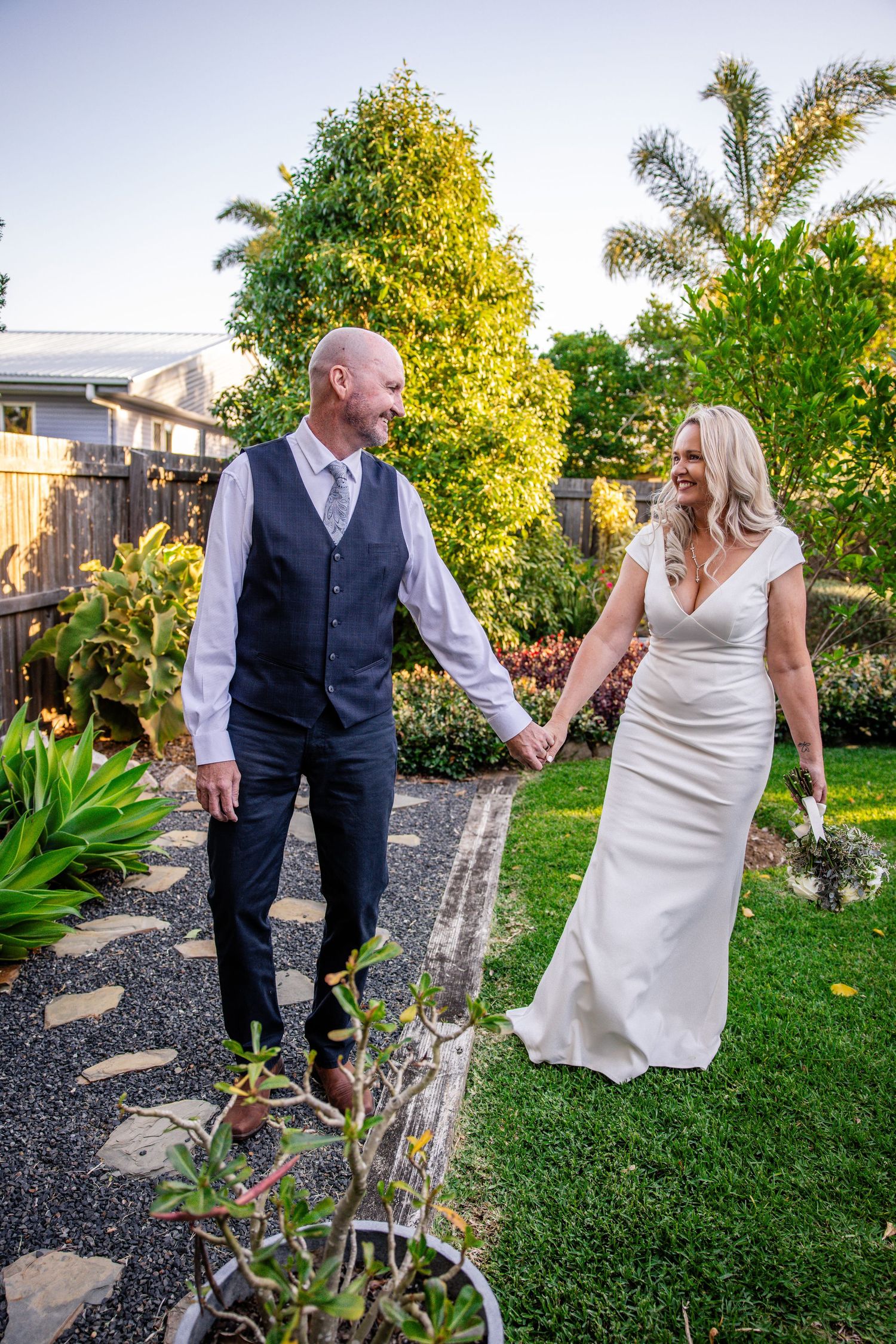 Couple in wedding attire holding hands and smiling in a lush garden setting with palm trees and landscaping.