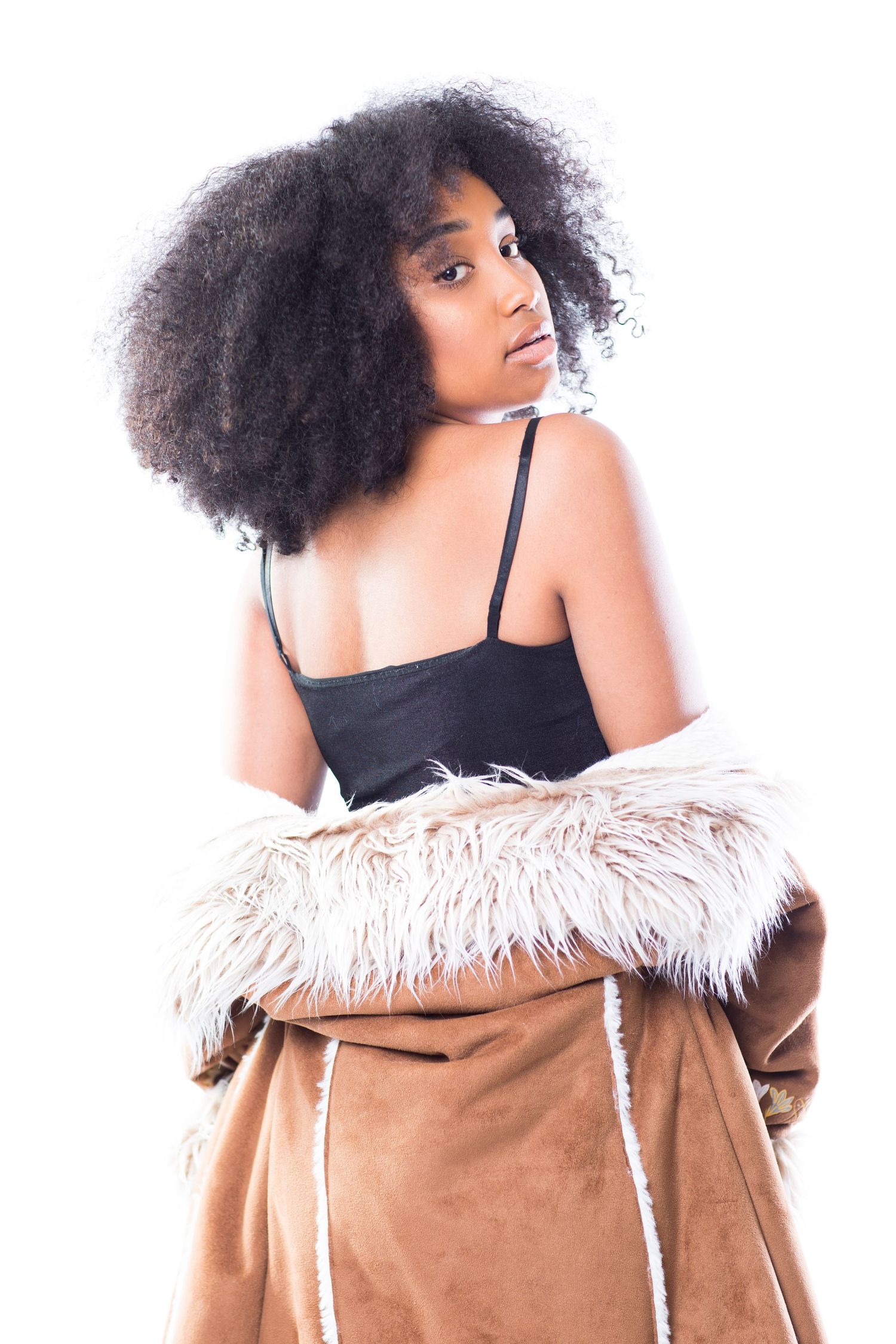 A stylish portrait of someone wearing a black tank top and brown fur-lined coat against a white background.