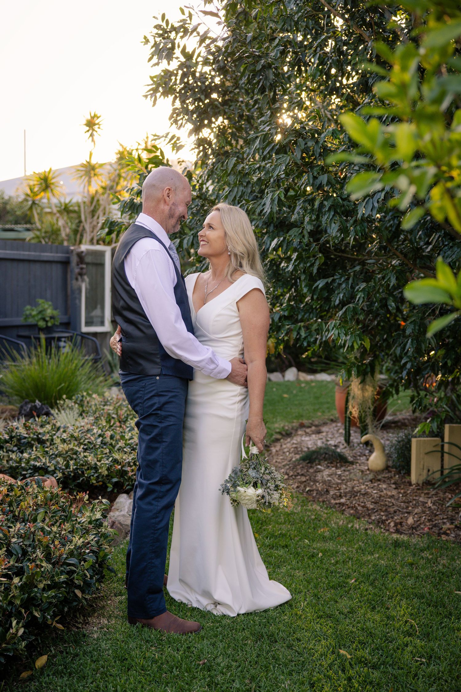 A romantic outdoor garden wedding moment with a couple embracing among lush greenery at sunset.