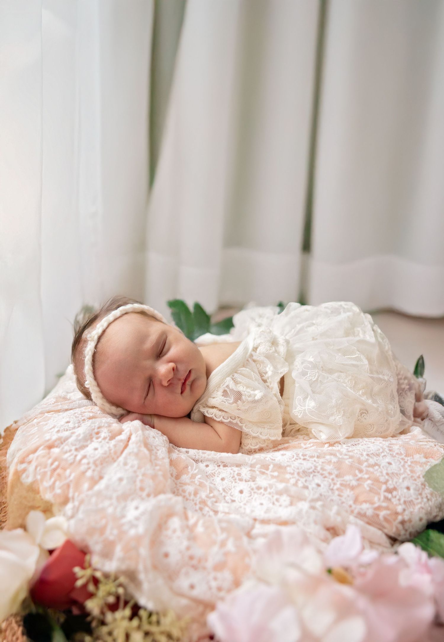 Sleeping newborn baby in ivory lace gown and headband resting on floral arrangement for professional portrait.