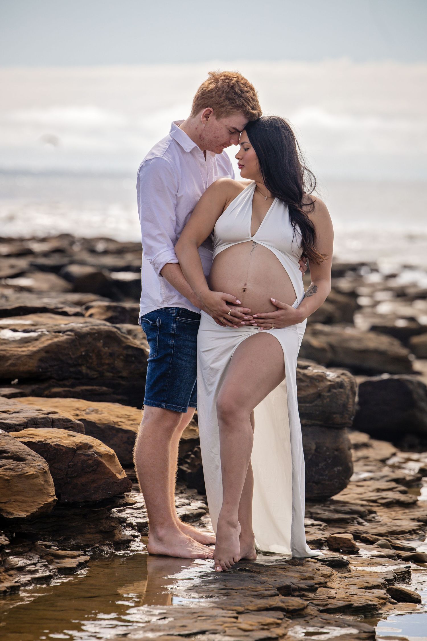 A romantic maternity photo shoot on a rocky beach at sunset shows a couple embracing near the ocean.