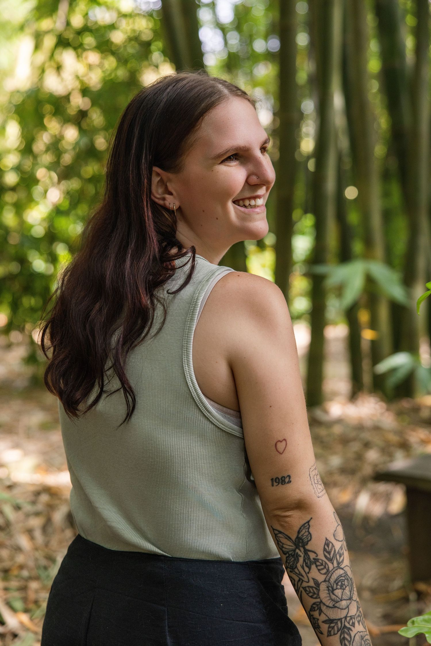 A person with tattoos and dark hair laughs joyfully in a sunlit forest setting wearing a sleeveless top.