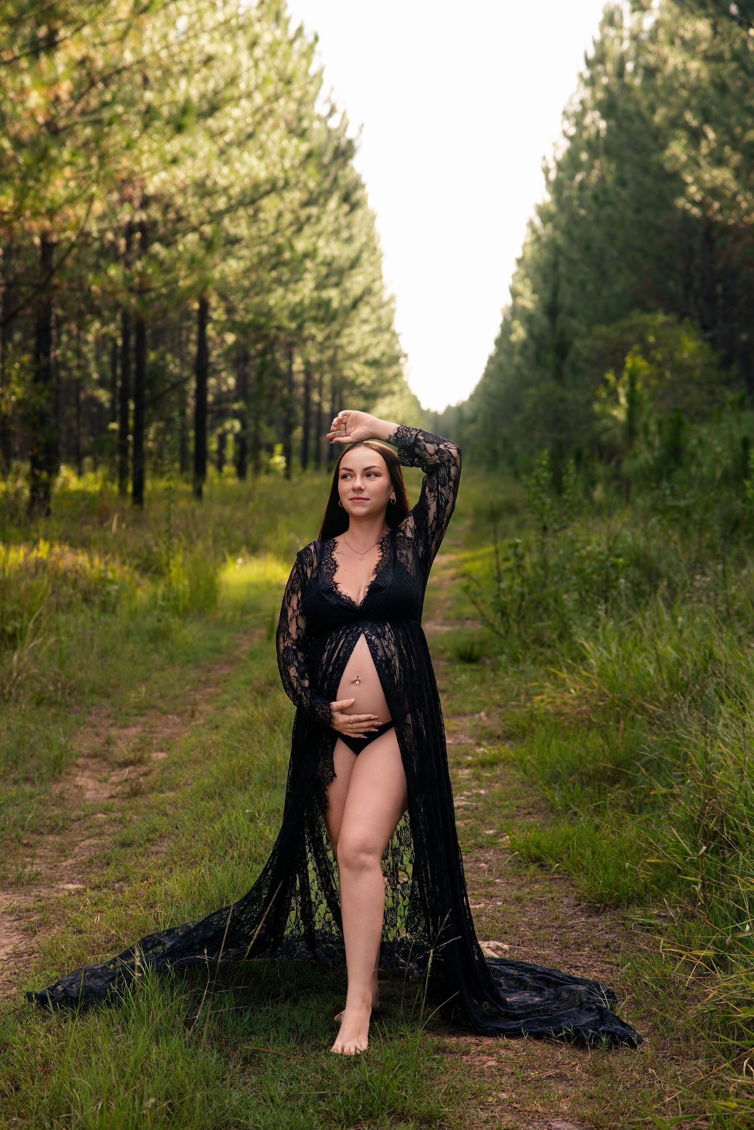 Pregnant woman in black lace robe and lingerie posing barefoot on a forest path surrounded by tall pine trees.