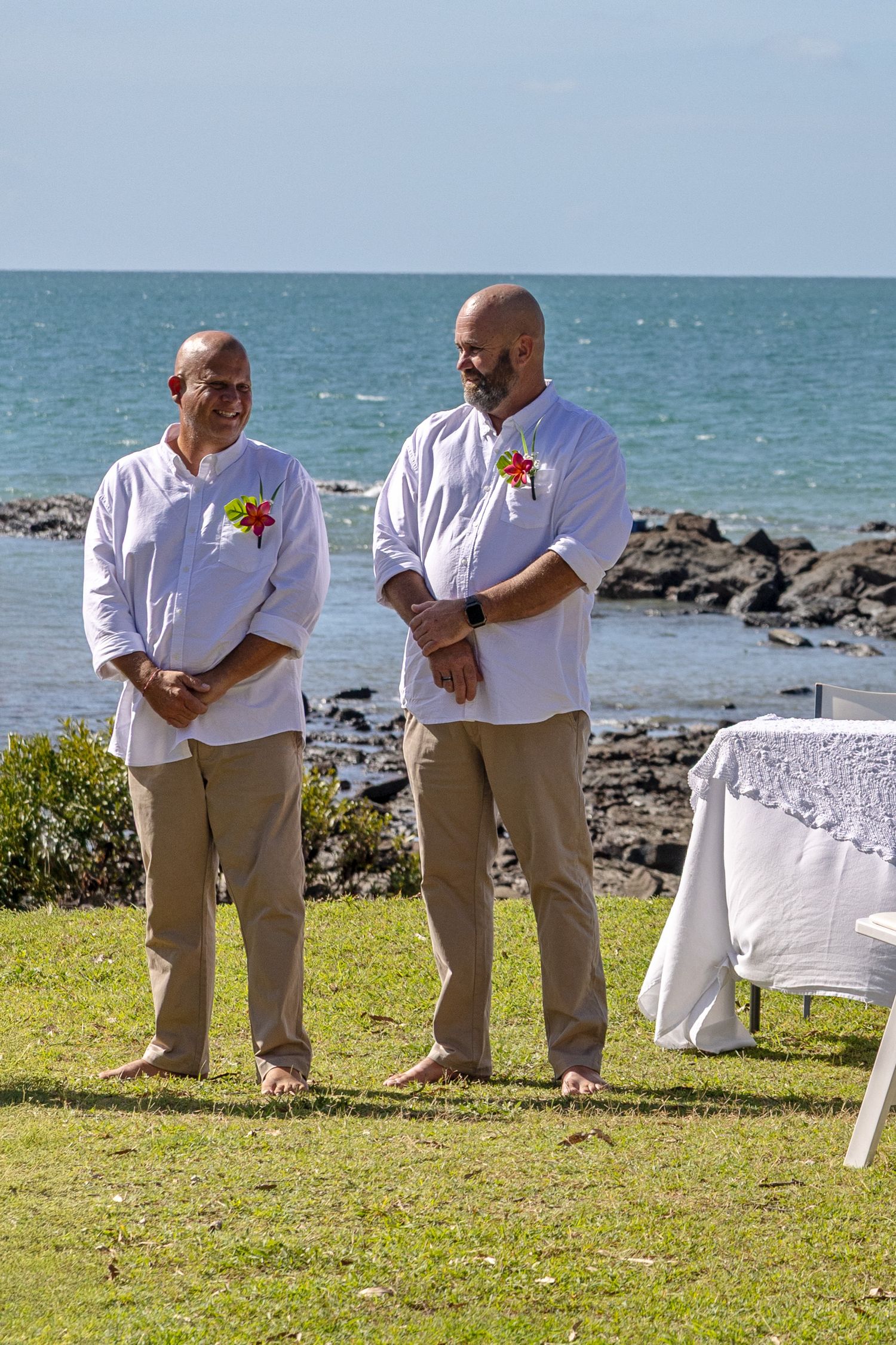 Two people in matching white shirts and khaki pants stand on grass by the ocean during an outdoor ceremony.