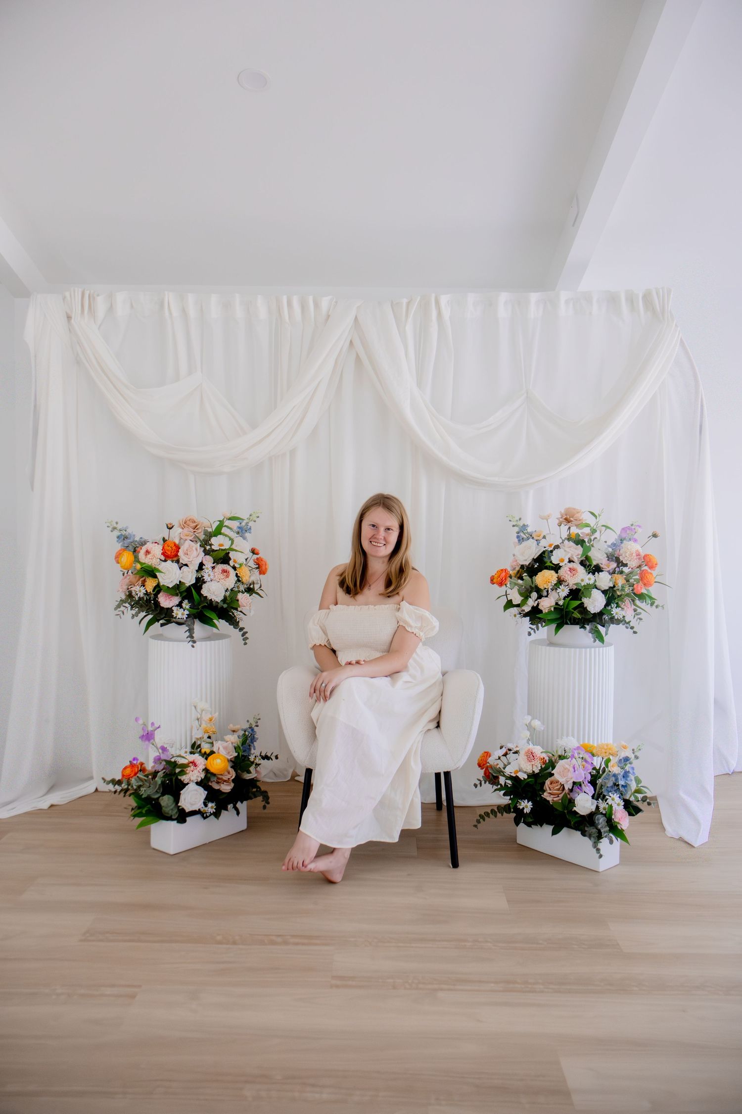 Woman in white dress sits on chair surrounded by colorful floral arrangements against white draped backdrop.