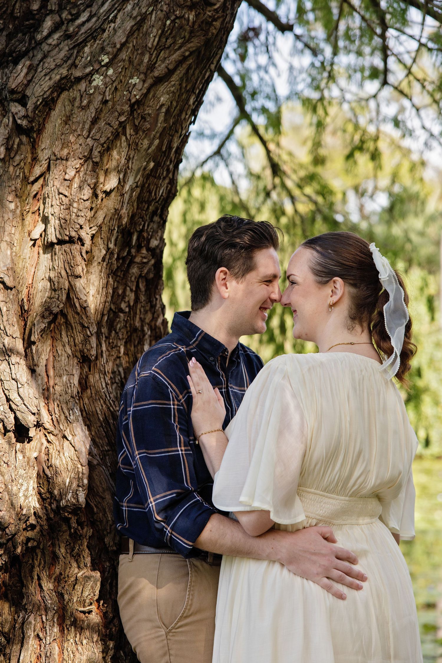 Couple in romantic embrace near a large tree, woman in white dress with veil, man in plaid shirt, outdoor park setting.