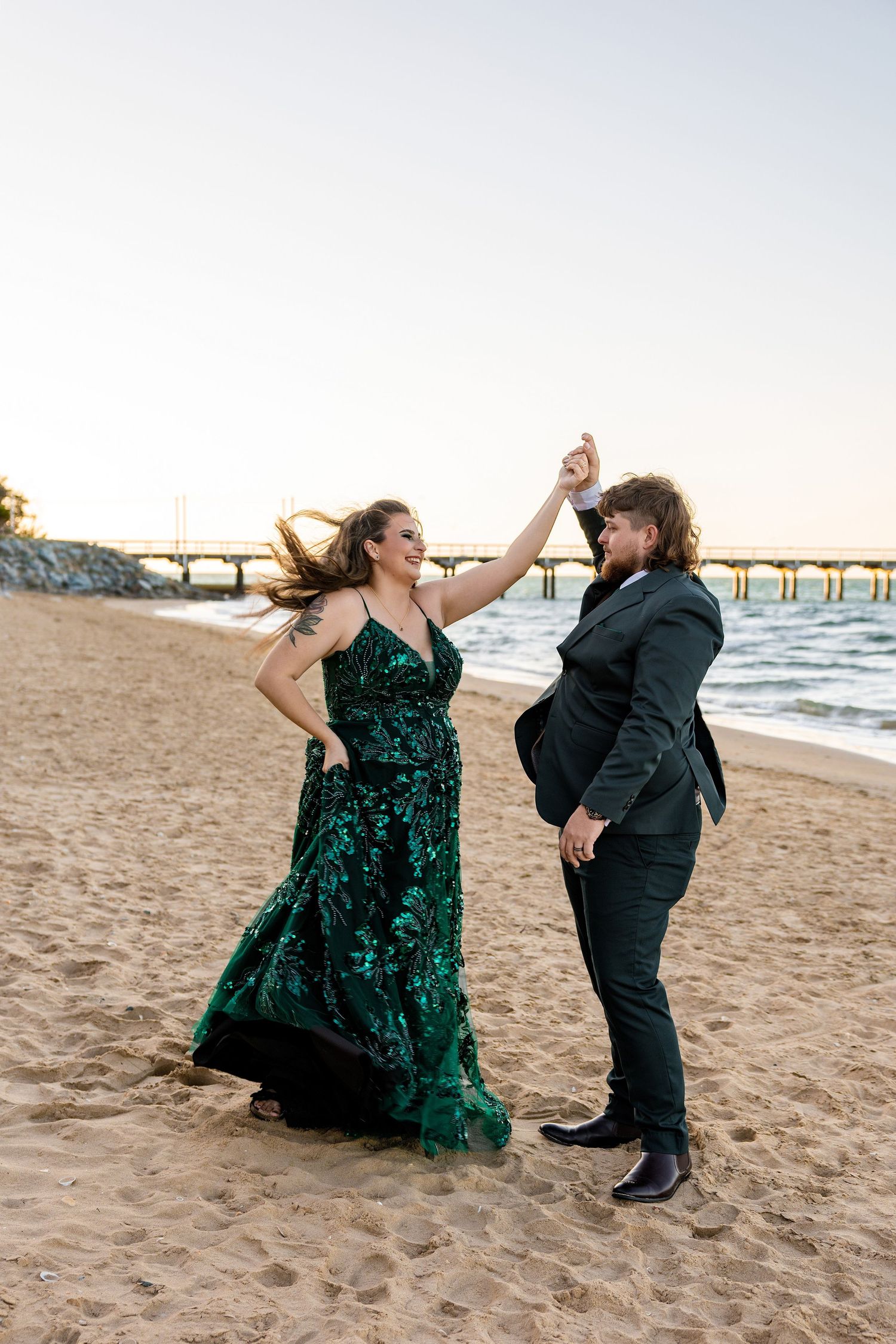 Couple dancing on beach at sunset, woman in green sequin gown, man in dark suit, pier in background.