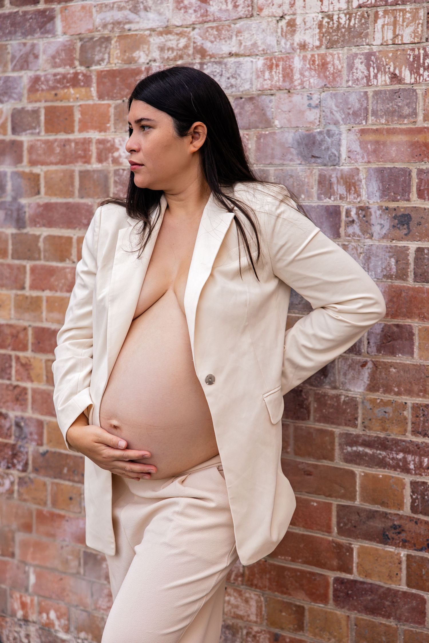 Expecting mother in cream suit posing against brick wall in maternity photoshoot.
