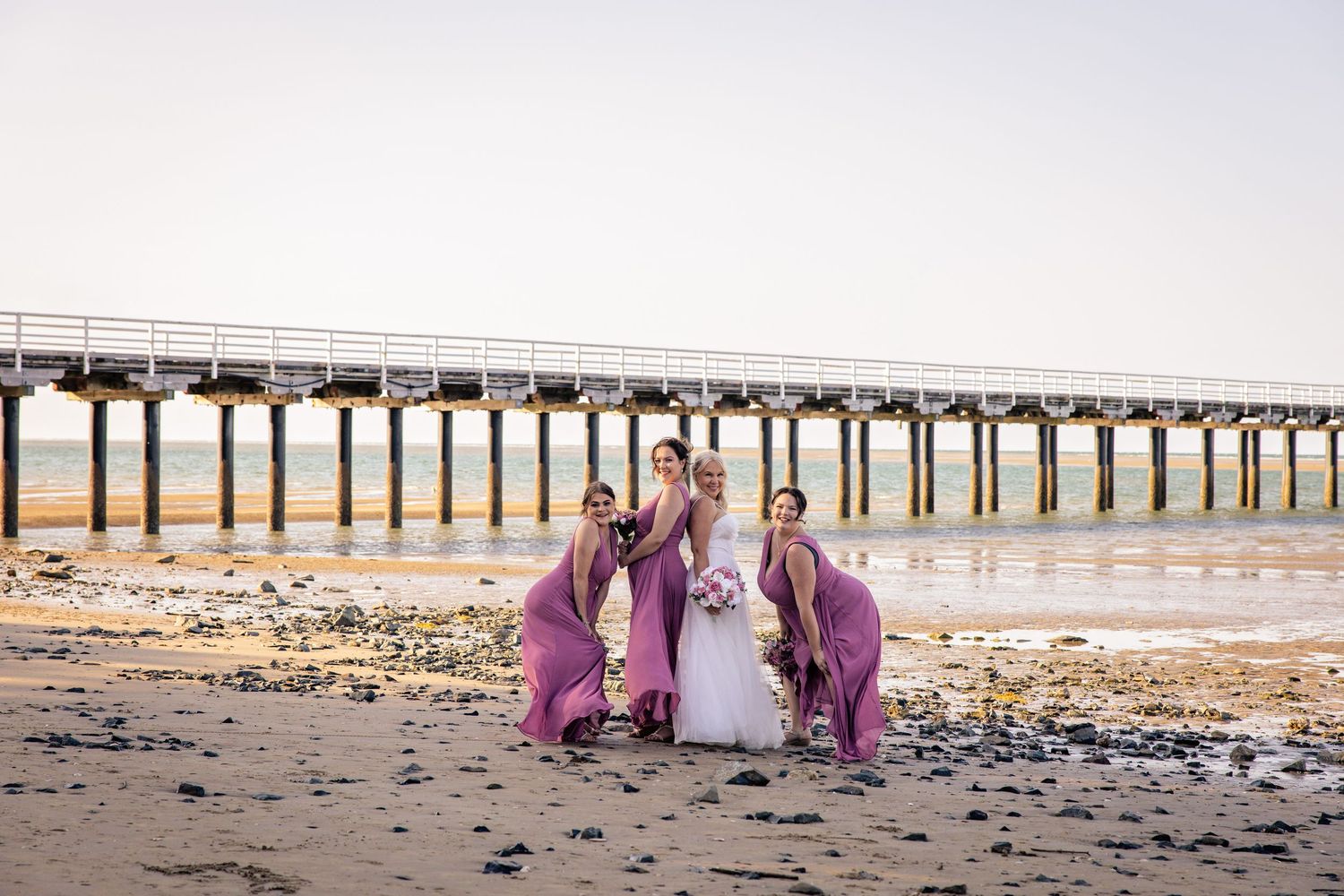 Wedding party in white and purple attire walking along sandy beach with long pier in background.