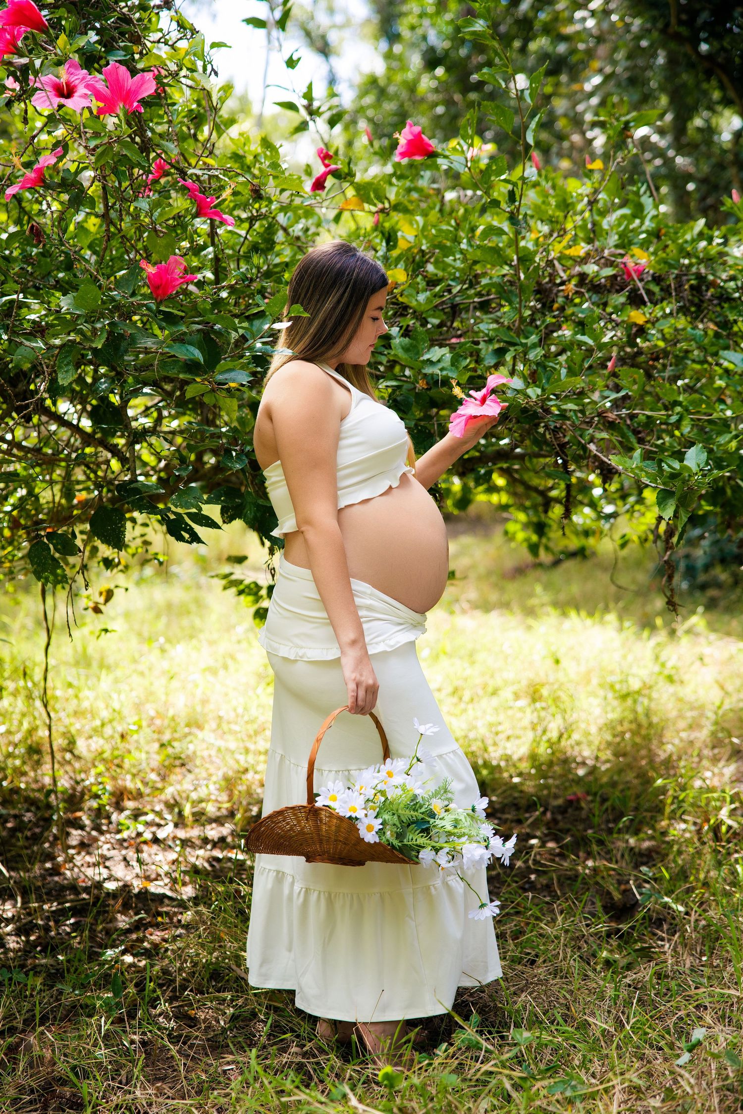 Pregnant woman in white dress holding a wicker basket with flowers, touching pink blooms in a lush garden.