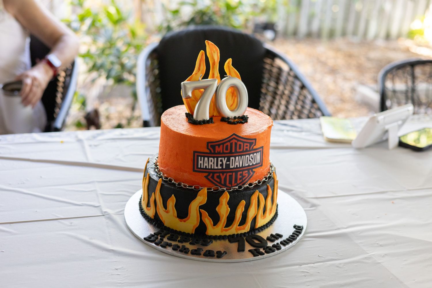 A two-tier Harley-Davidson themed 70th birthday cake decorated with orange flames on a white table outdoors.