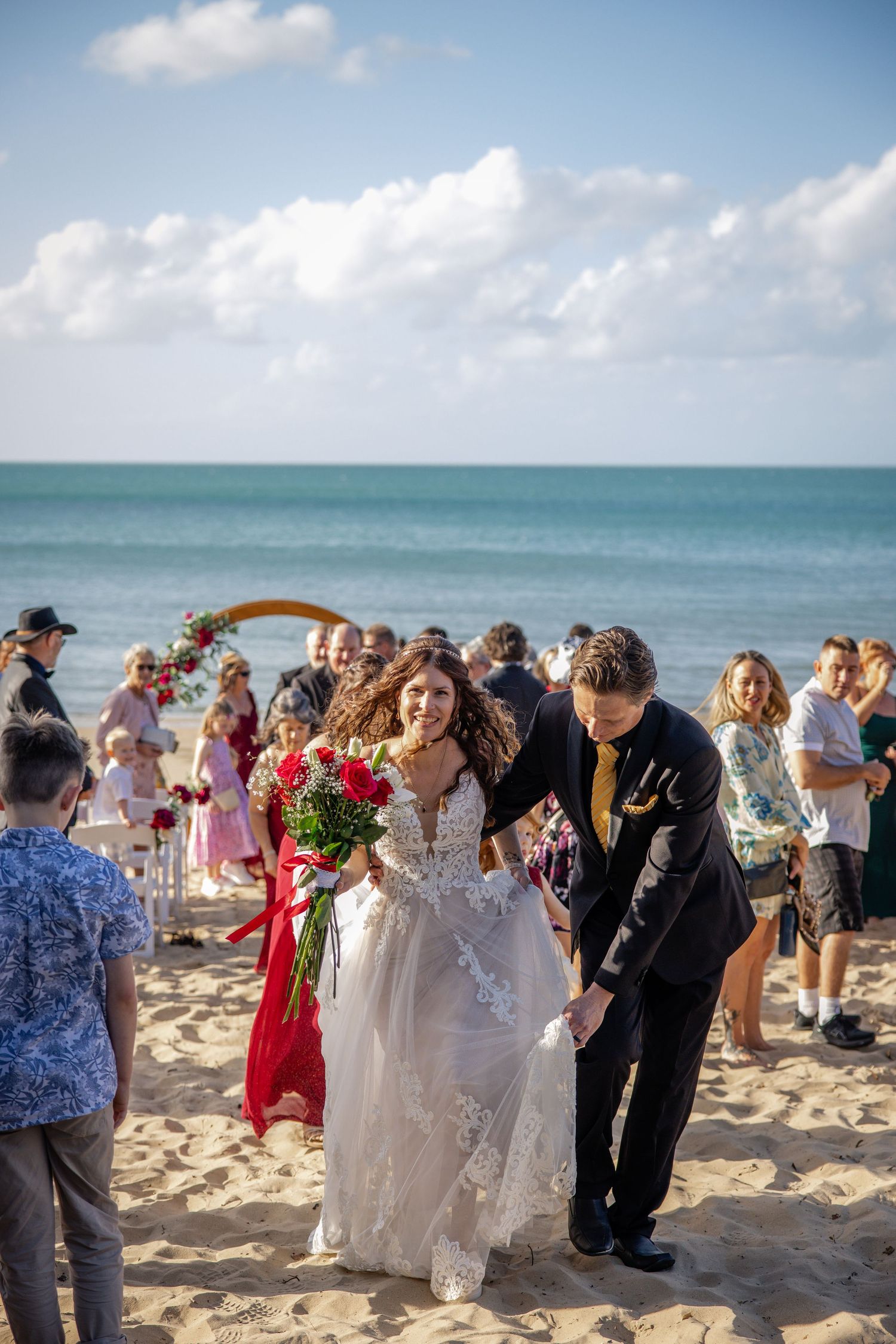 Wedding guests gather on a tropical beach under blue skies with turquoise waters for a seaside ceremony.