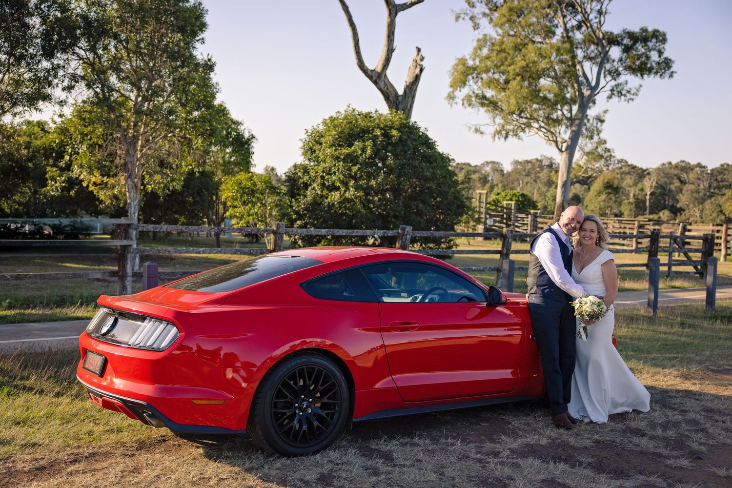 A red Ford Mustang GT parked in a scenic outdoor setting during sunset with a couple posing beside it.