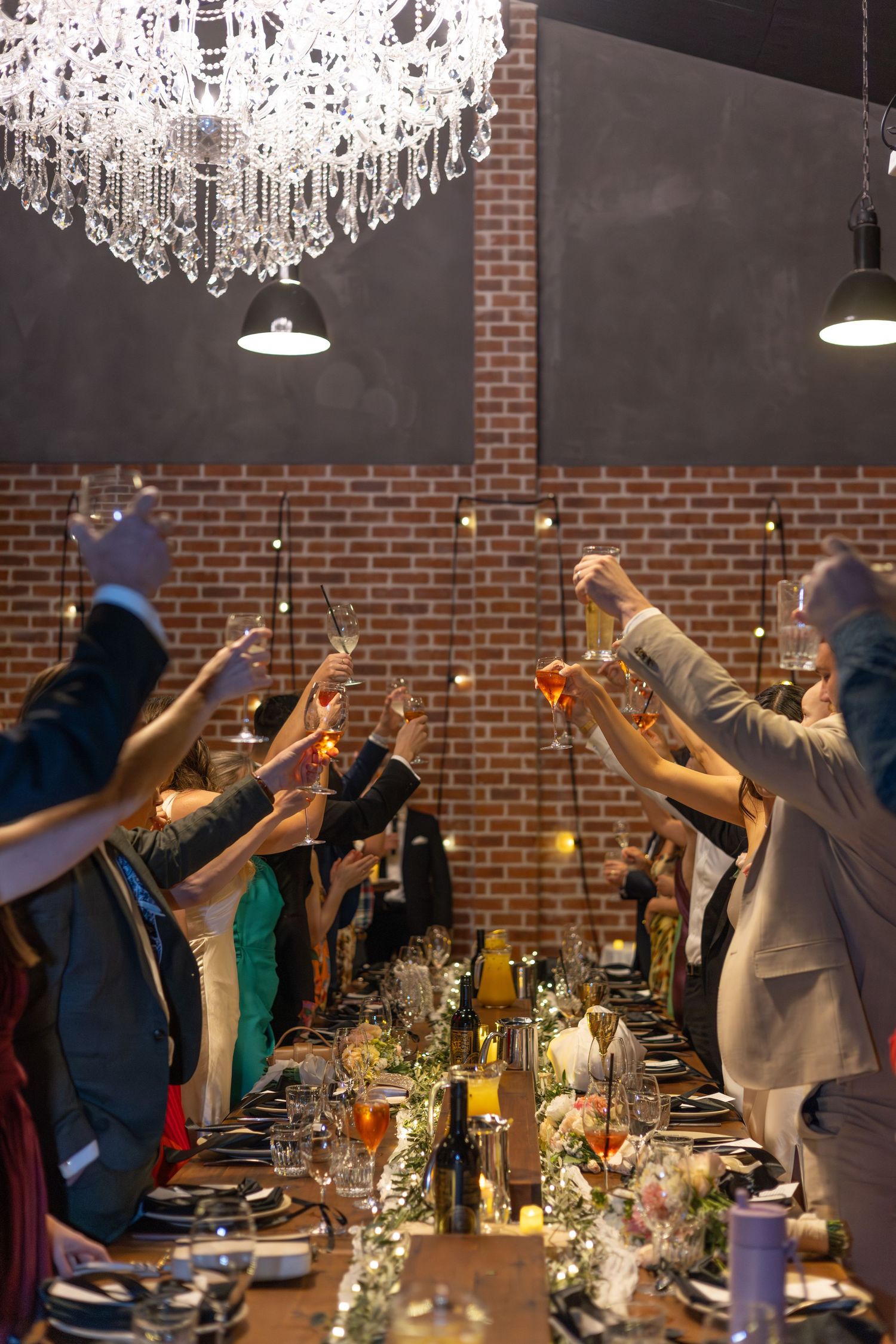 Dinner guests raise their glasses in a toast under a crystal chandelier at an elegant brick-walled restaurant.