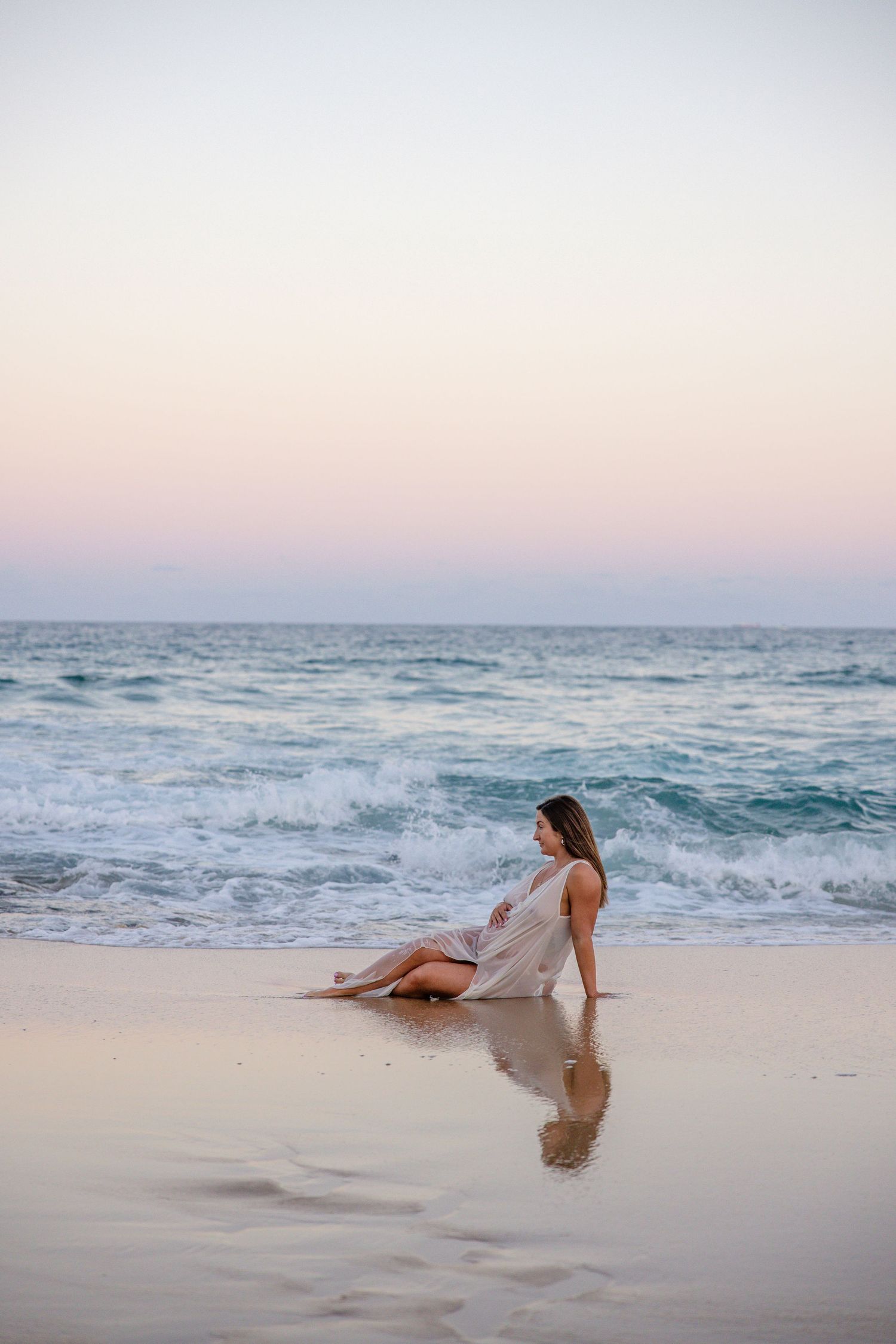 A person in white dress sits peacefully on a sandy beach at sunset as ocean waves roll in.