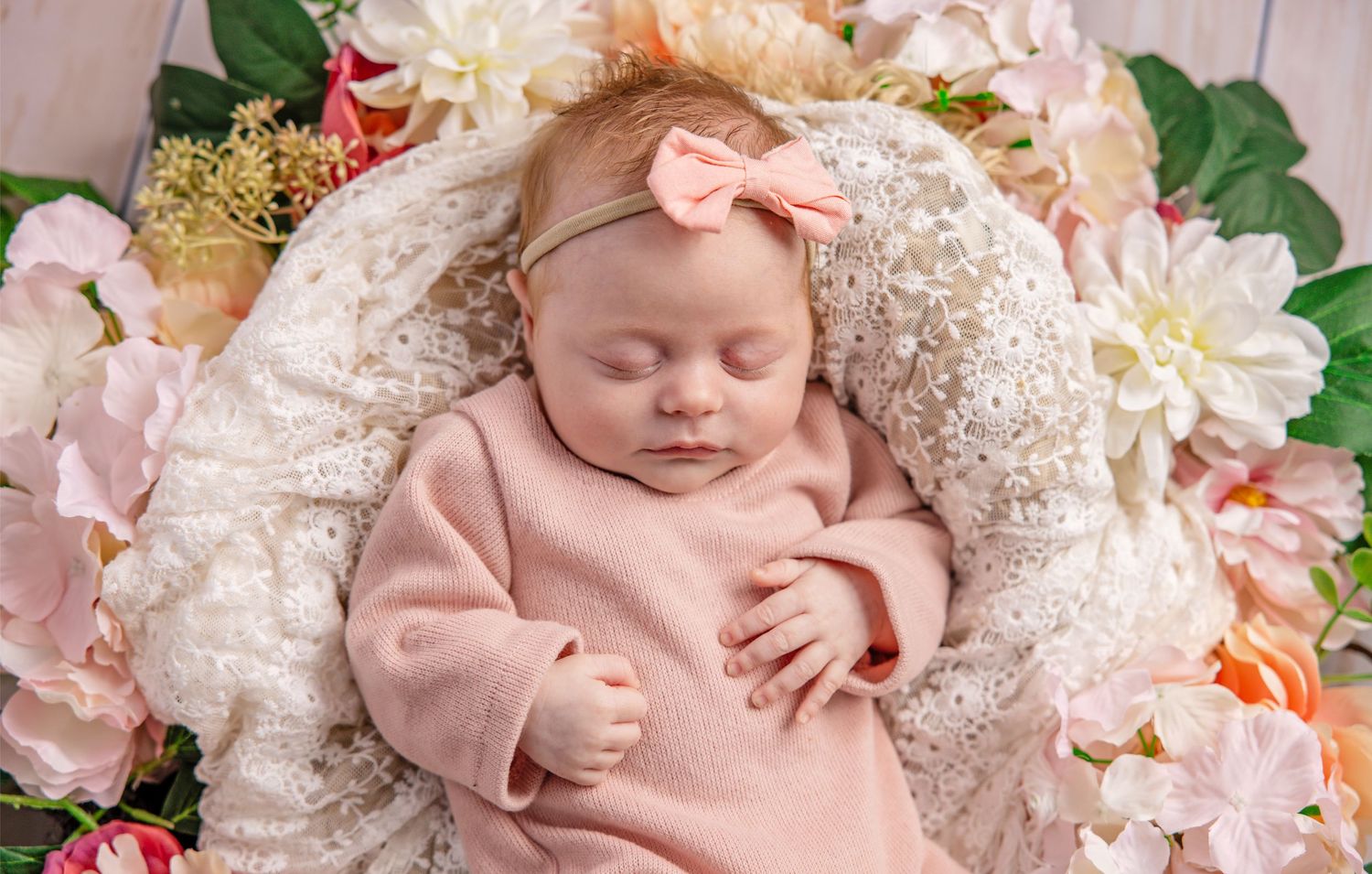 Newborn baby wearing pink outfit lies peacefully on white lace fabric surrounded by delicate floral decorations.
