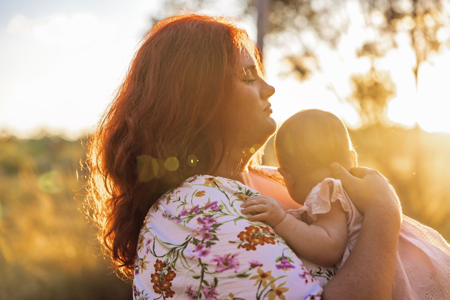 A mother with red hair holds her baby in golden sunset light outdoors.