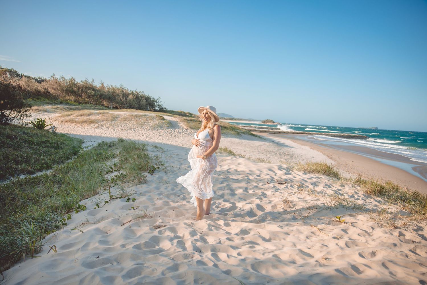 Pregnant woman in white dress and hat walking on sandy beach path with ocean waves and blue sky.