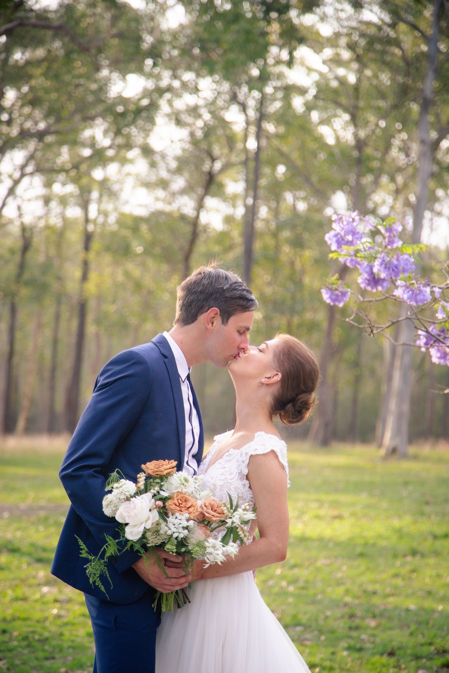 Romantic wedding couple kissing in a sunlit forest with bouquet of flowers.