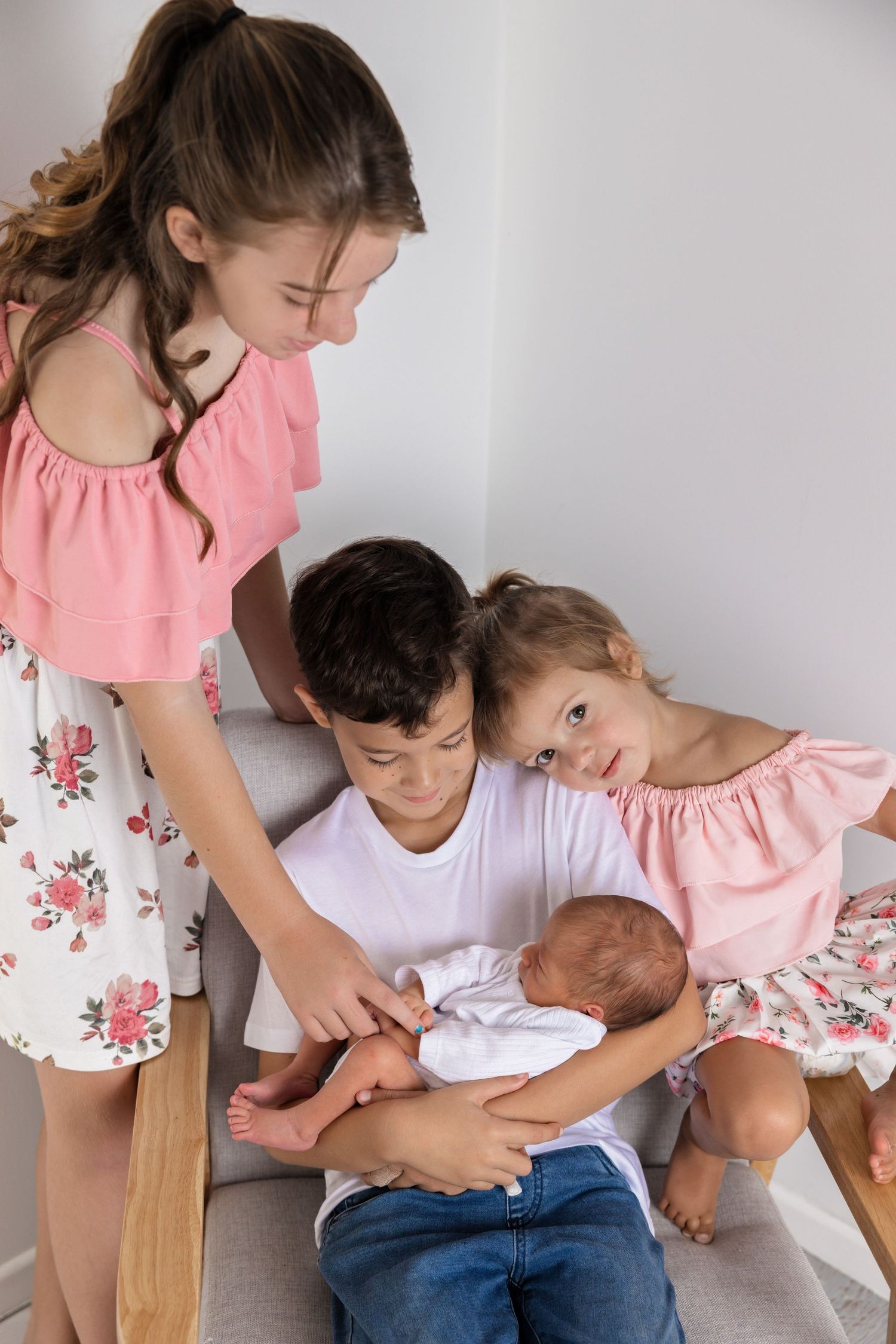 Children in pink and white outfits looking adoringly at a newborn baby sitting together on a wooden bench.