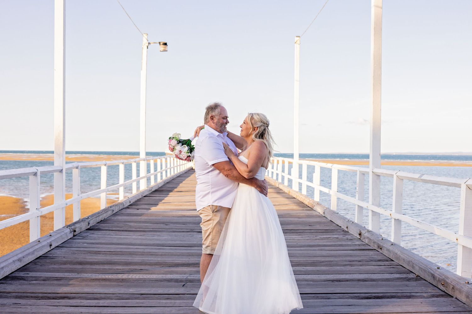 A couple shares a romantic moment on a wooden pier overlooking a serene waterfront at sunset.