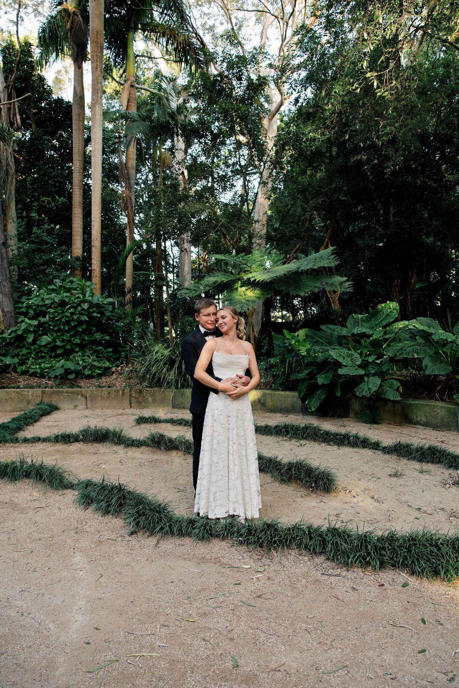 Bride and groom embracing in a lush garden maze with tropical greenery and palm trees backdrop.
