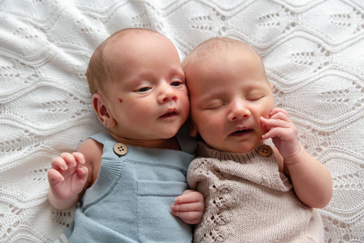 Two newborn twins sleeping peacefully side by side on a white knit blanket, dressed in blue and beige outfits.