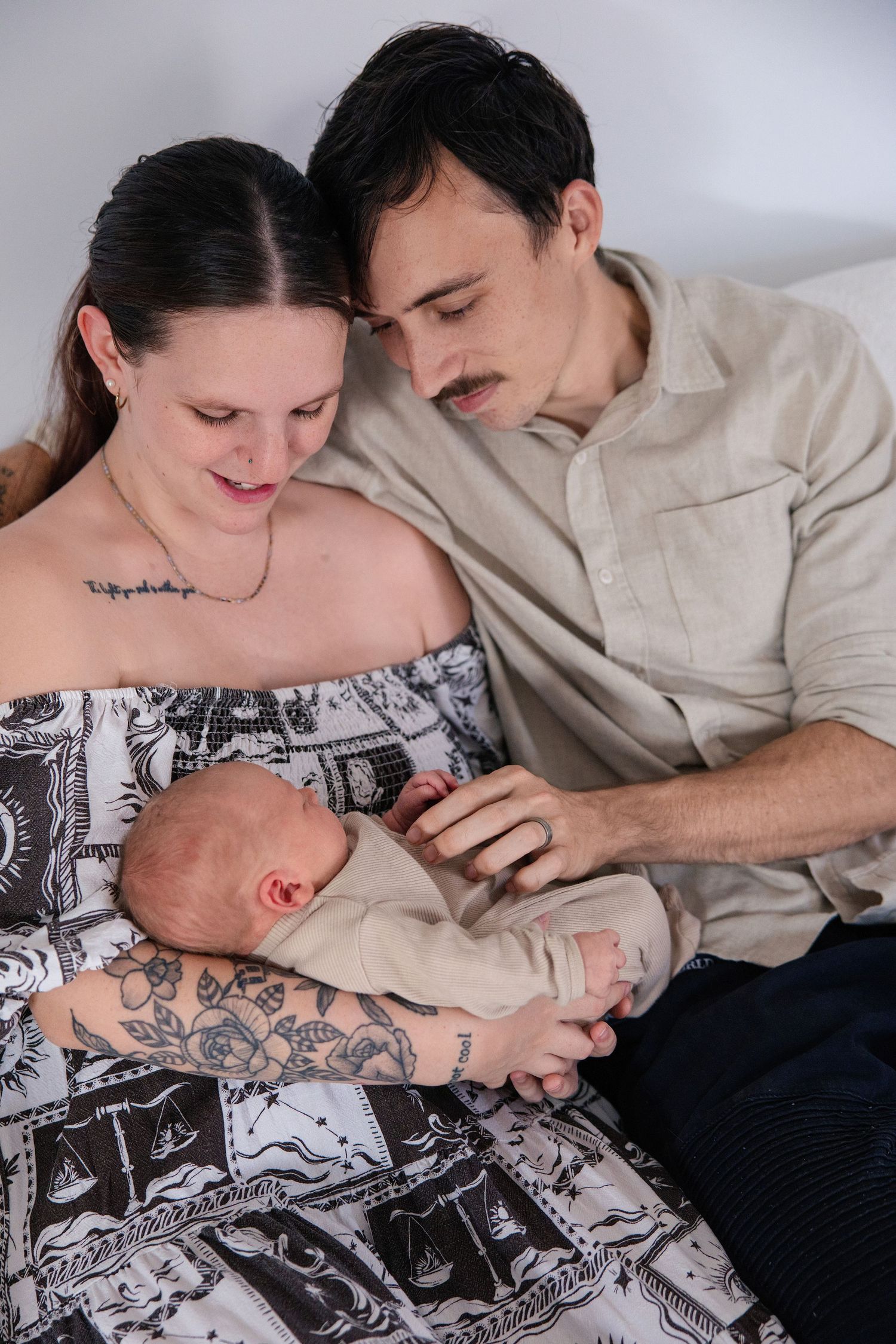 A couple with tattoos tenderly gazes at their newborn baby, sitting together in a warm and intimate family portrait.