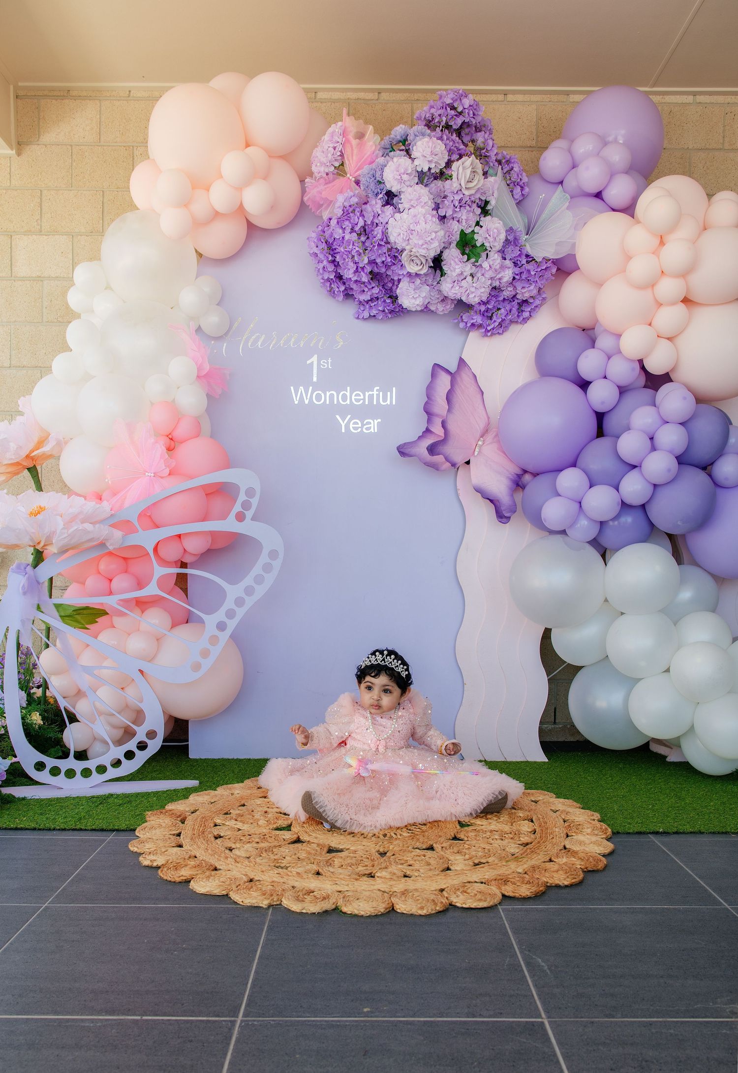 Baby girl in pink dress sits on rug at 1st birthday party with purple and pink balloon arch backdrop.