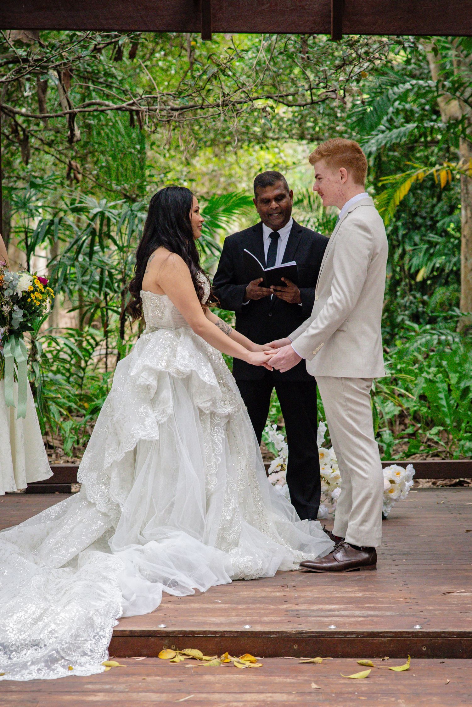 Wedding ceremony taking place outdoors on a wooden deck surrounded by lush greenery and trees.