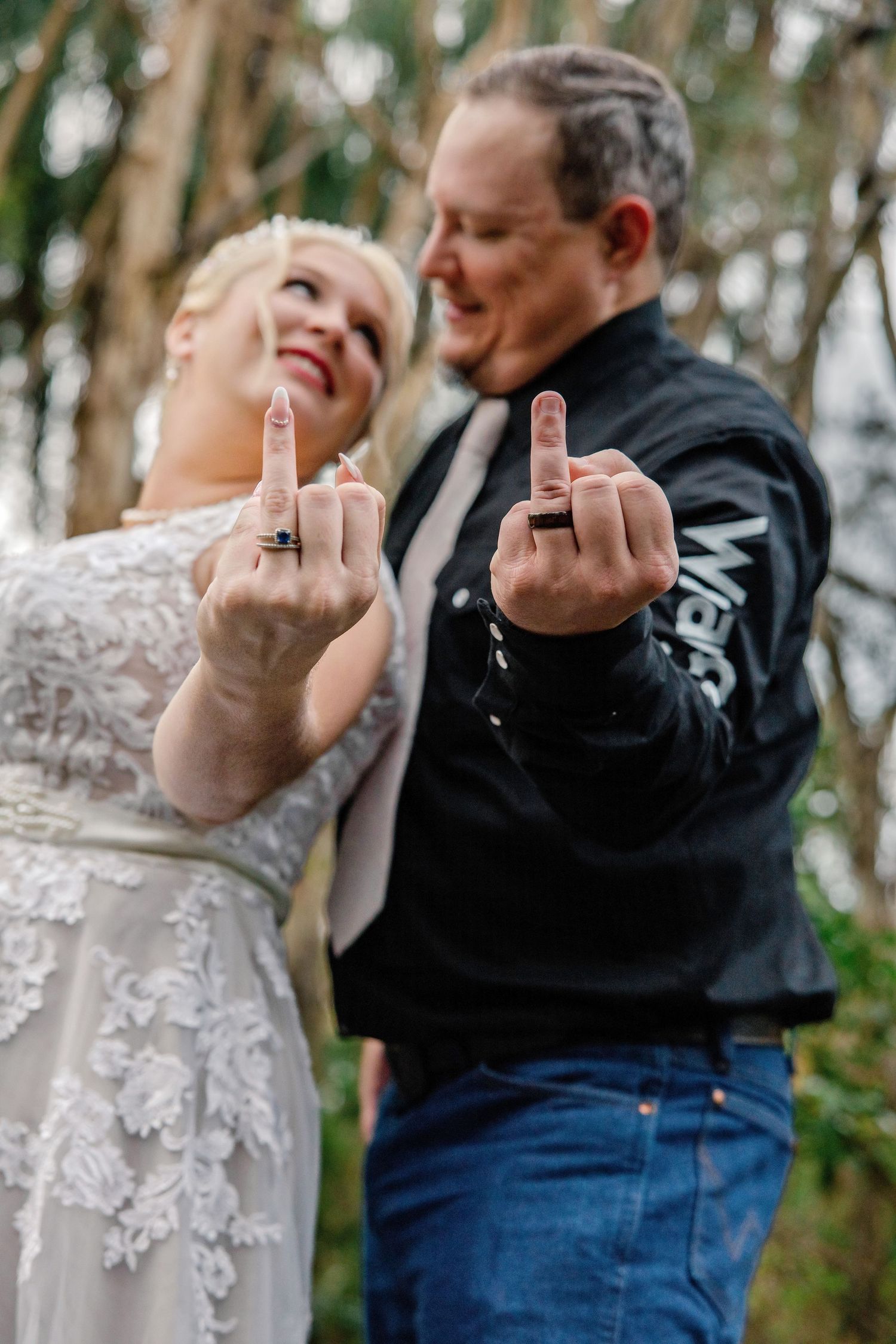 Newlywed couple in wedding attire showing off rings on middle fingers, smiling outdoors near trees.