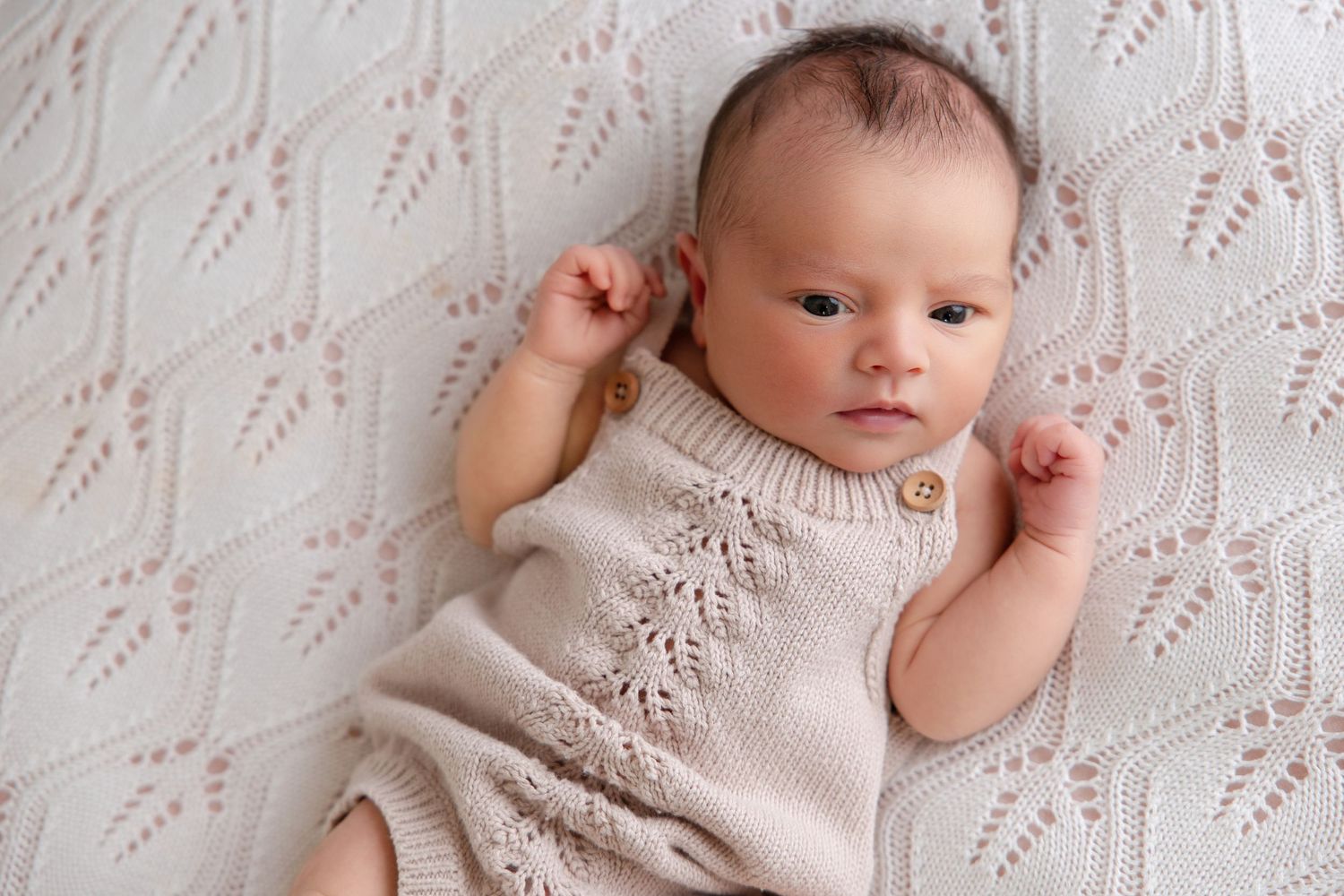Newborn baby in knit romper lying on white textured blanket, alert with fists raised.