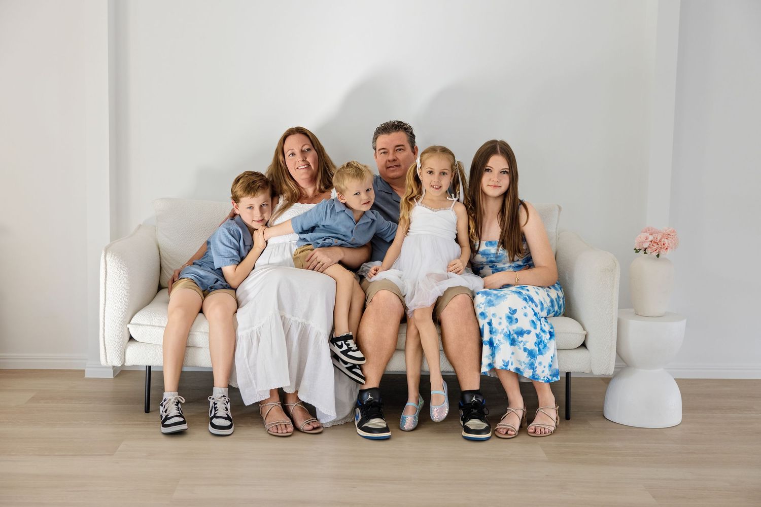 Family of six posing together on a white couch in a bright studio, smiling warmly for a professional portrait photo.