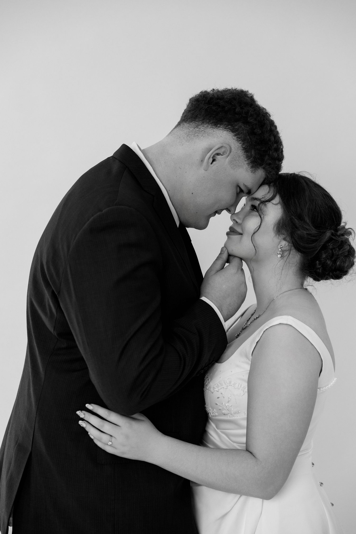 Black and white photo of a bride and groom sharing an intimate moment, touching foreheads and holding hands on their wedding day.