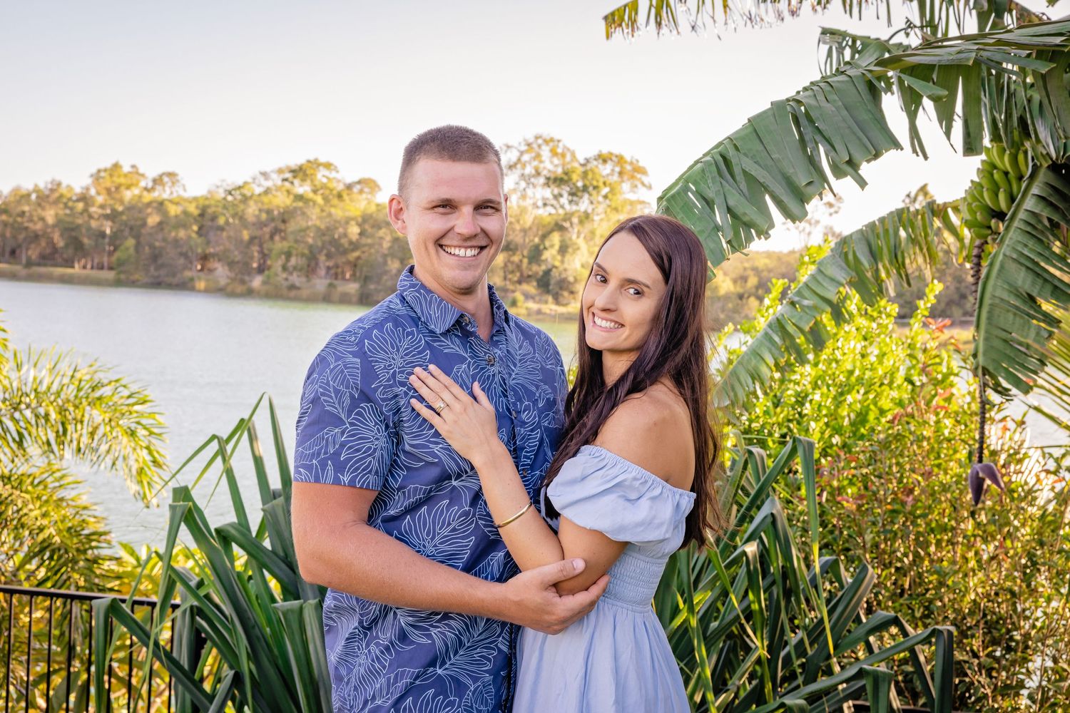 A romantic couple embracing near a tropical waterfront setting at sunset.