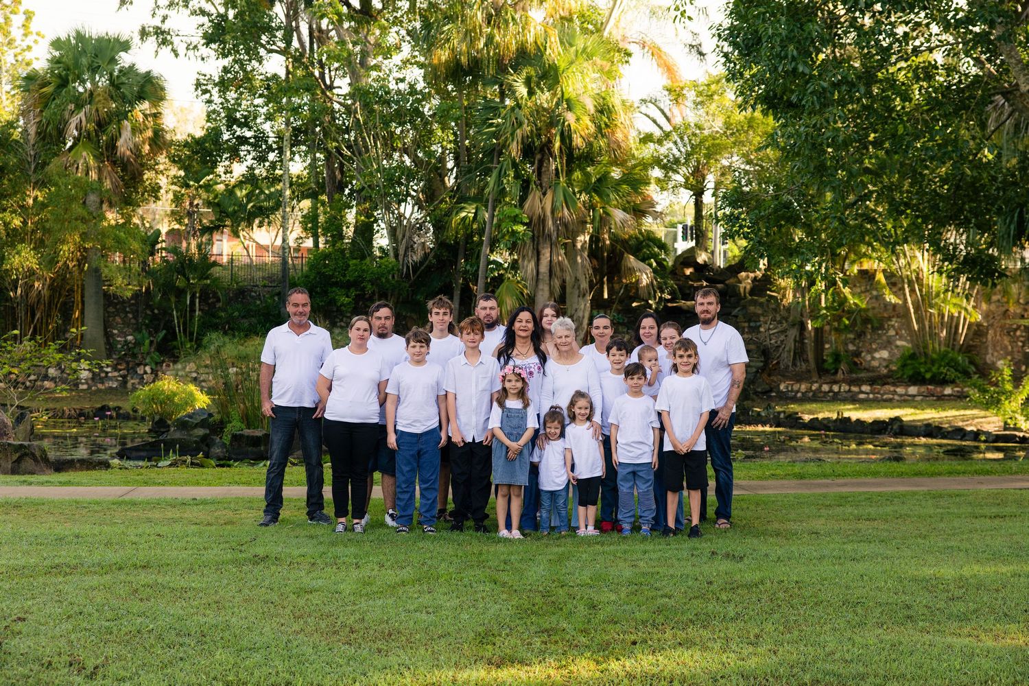 Large family group portrait outdoors on green lawn surrounded by lush tropical trees, all wearing white tops and jeans.