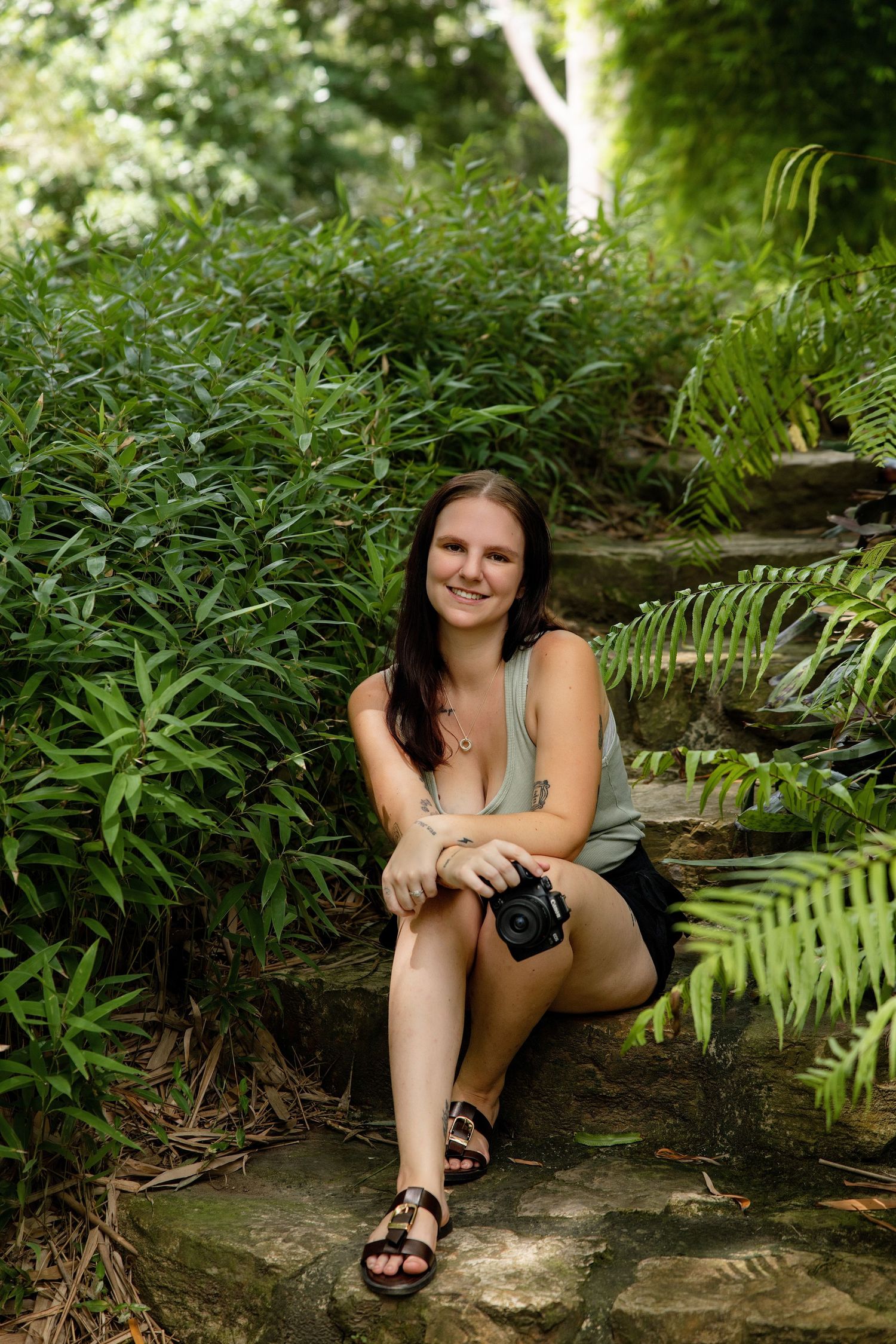 Young woman sitting on stone steps in a lush green garden, smiling and holding a camera, wearing a tank top and shorts.