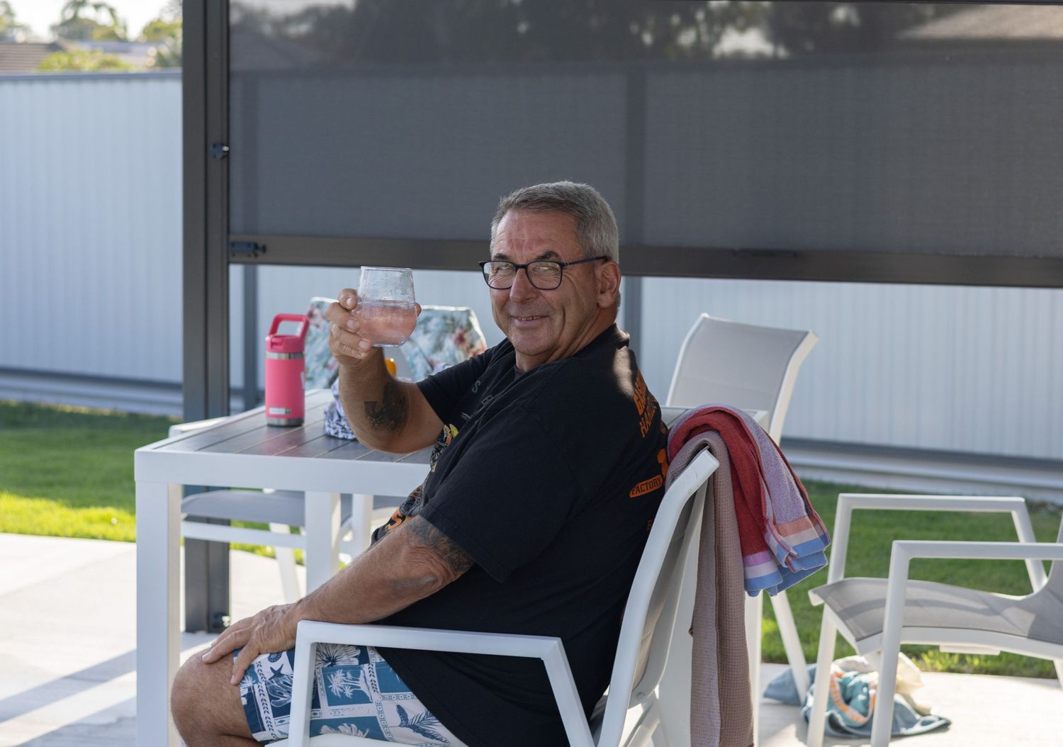 A person relaxes in a white Adirondack chair on a sunny porch while holding a beverage.