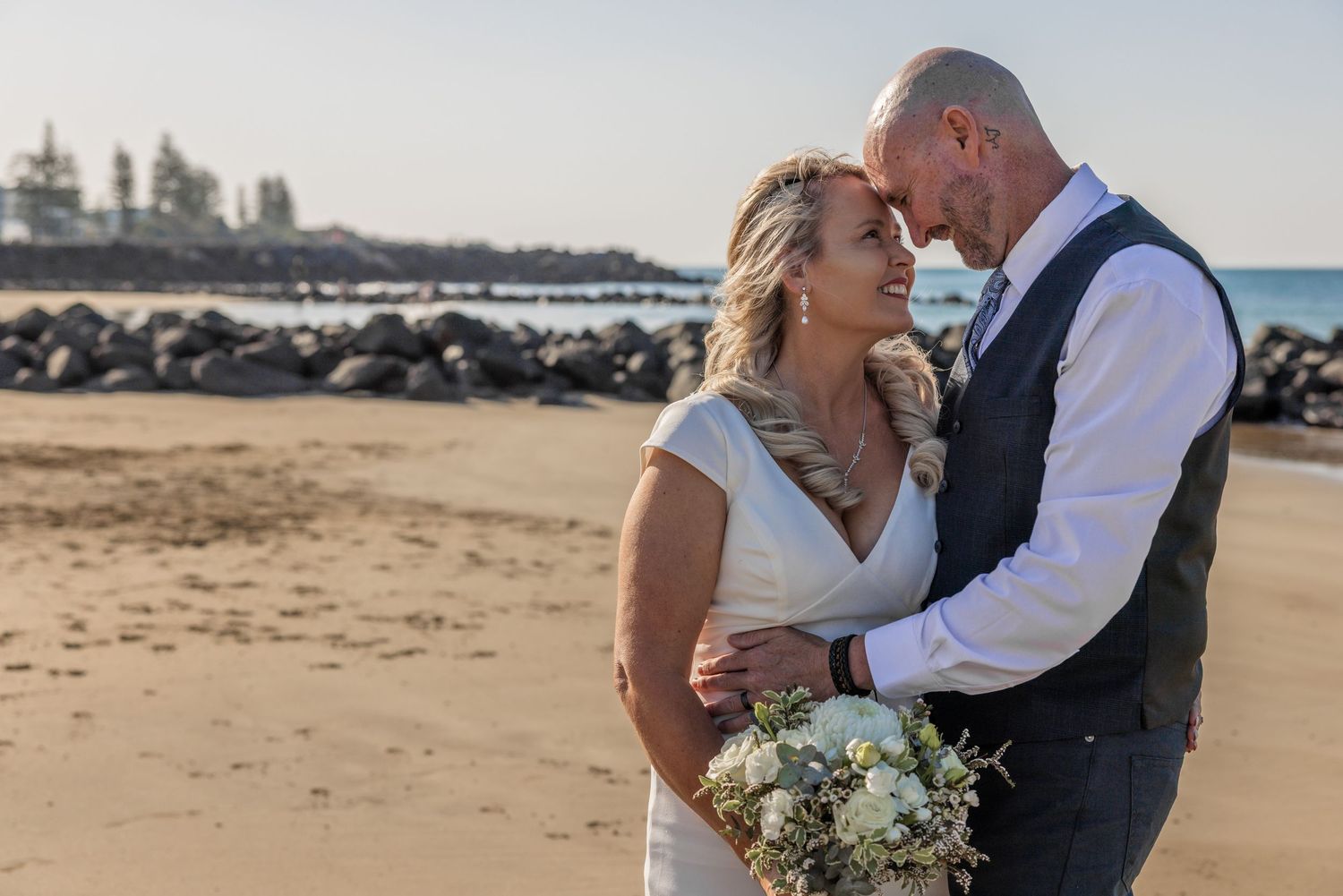 A romantic wedding couple embraces on a scenic beach with ocean waves and sand in the background during golden hour.