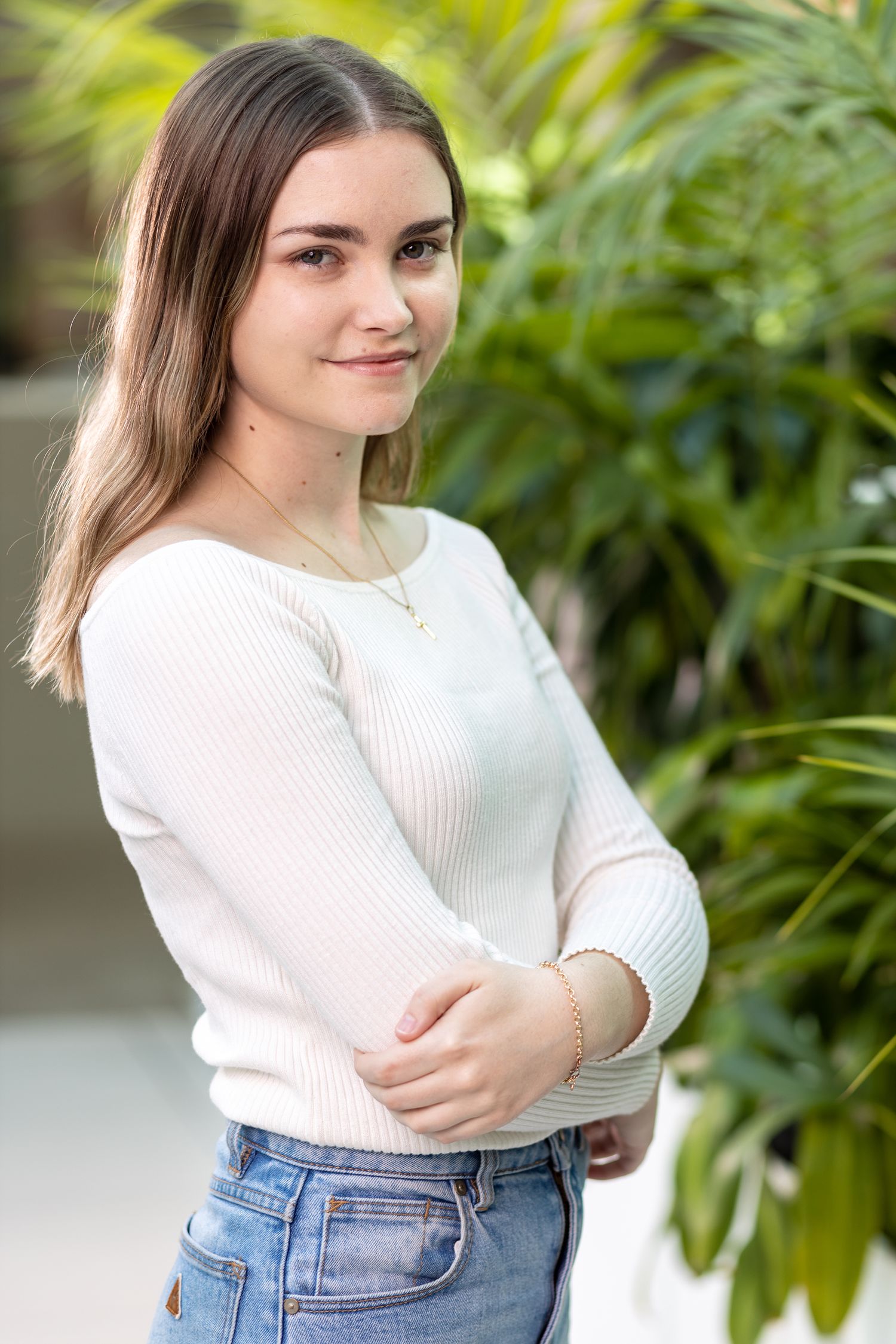 Young woman in white sweater and jeans poses against tropical foliage background.