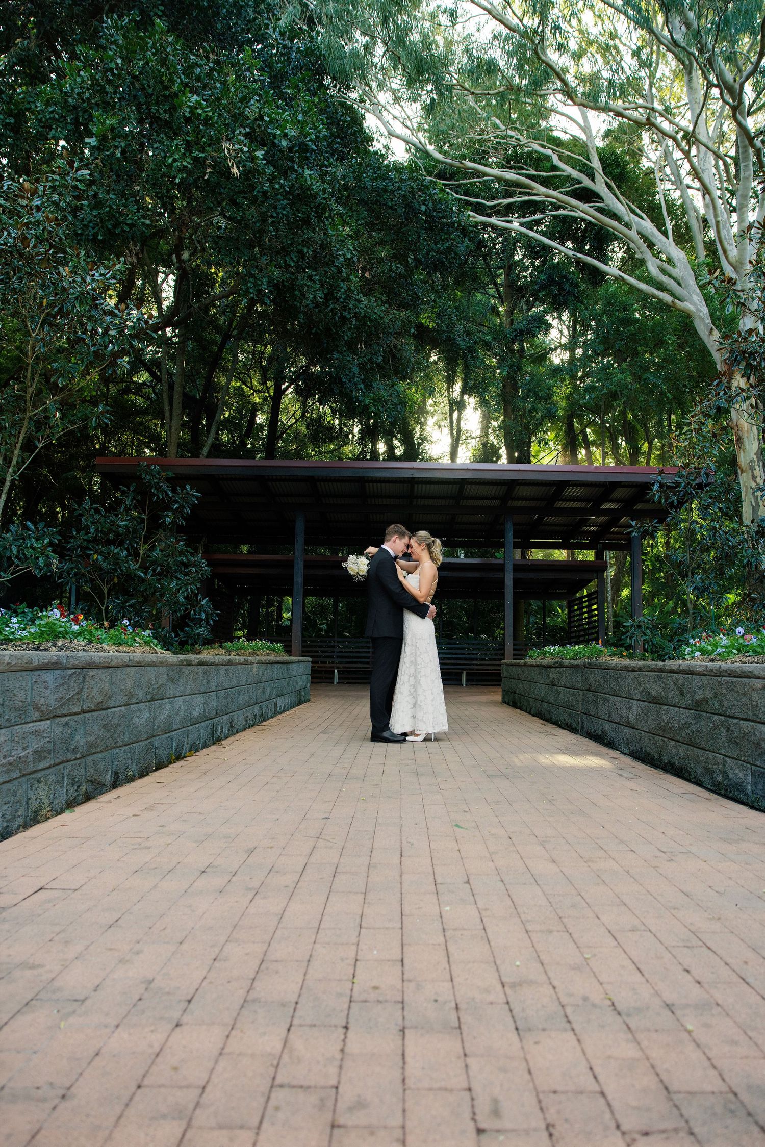 Bride and groom share a romantic kiss on a brick pathway surrounded by lush greenery at an outdoor wedding venue.