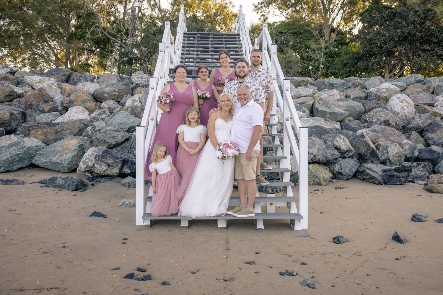 Wedding group poses on rocky beach with white arch and mauve dresses at sunset.
