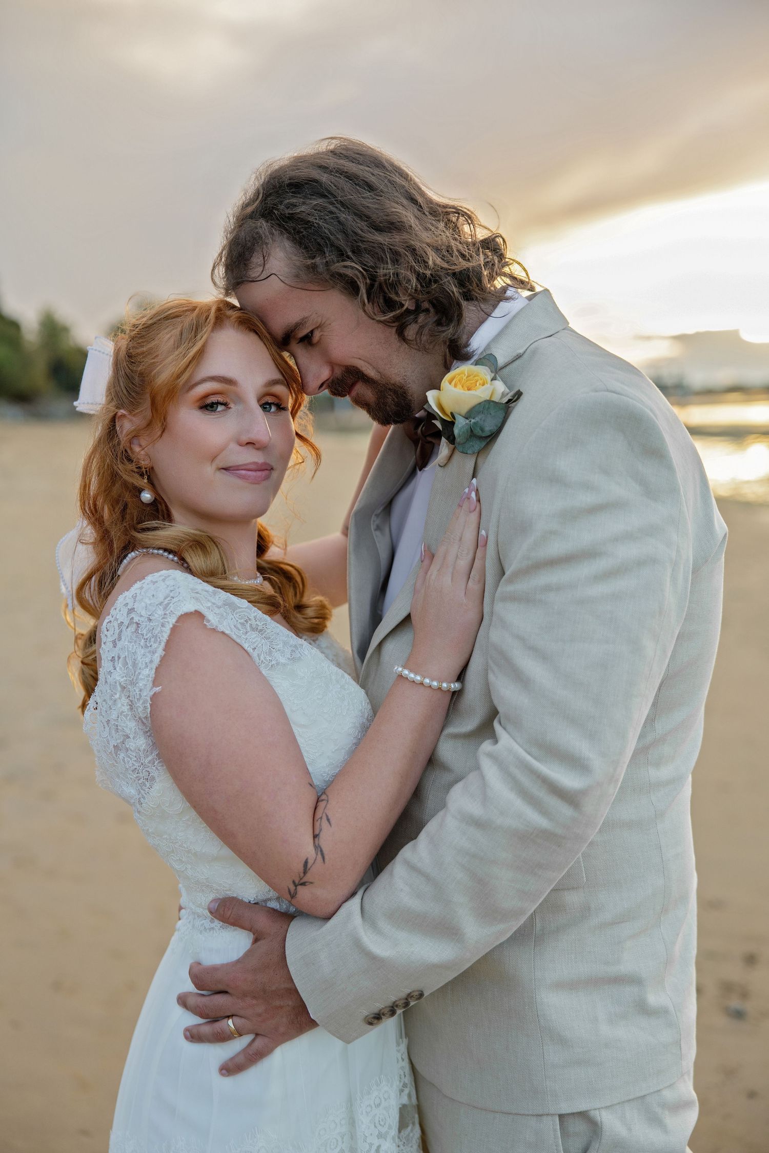 Bride and groom embrace on sandy beach at sunset, she in lace dress with pearl bracelet, he in beige suit with yellow boutonniere.
