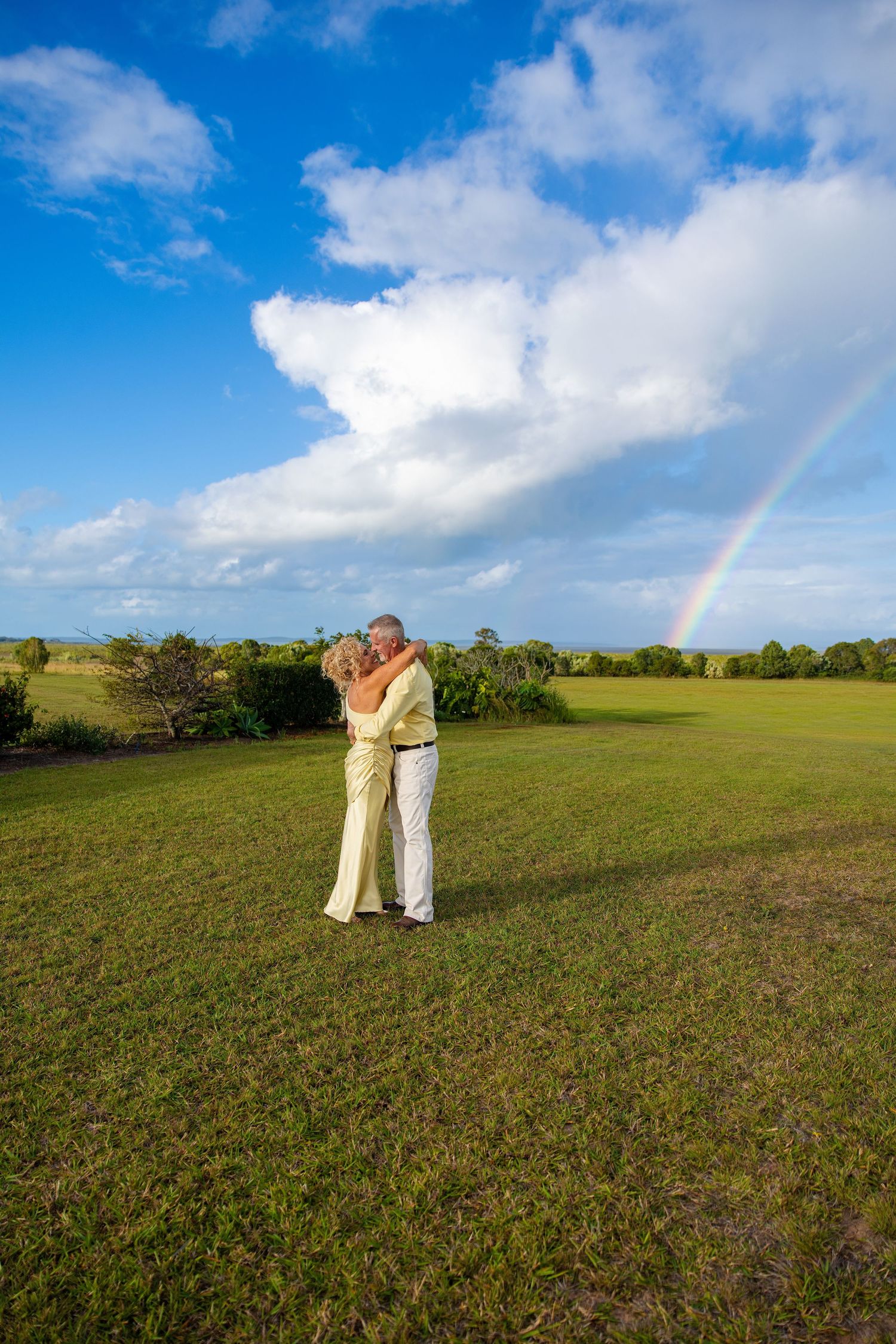 Couple embracing on green lawn with rainbow and blue sky in background.