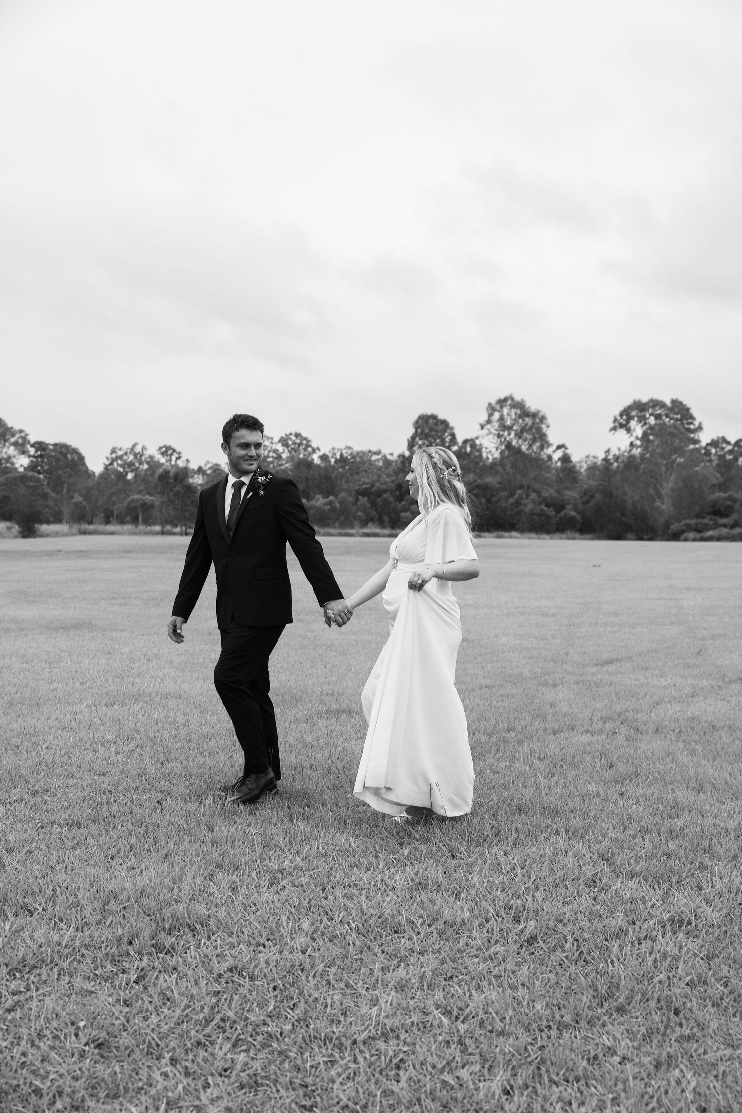 A bride and groom walk hand in hand across a grassy field in black and white photography.