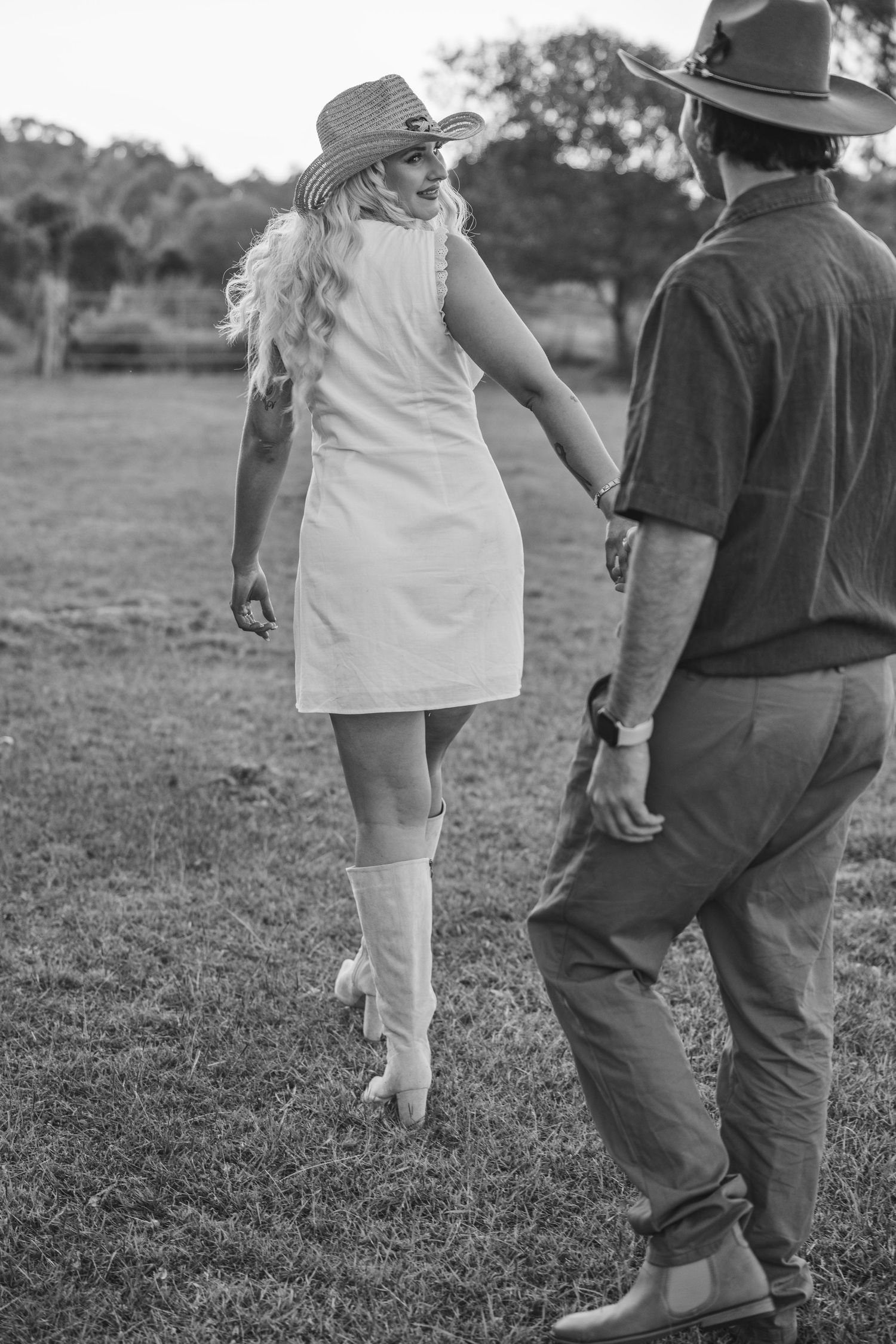 Black and white retro photo of a woman in a white dress and knee-high boots walking with a cowboy in a field.