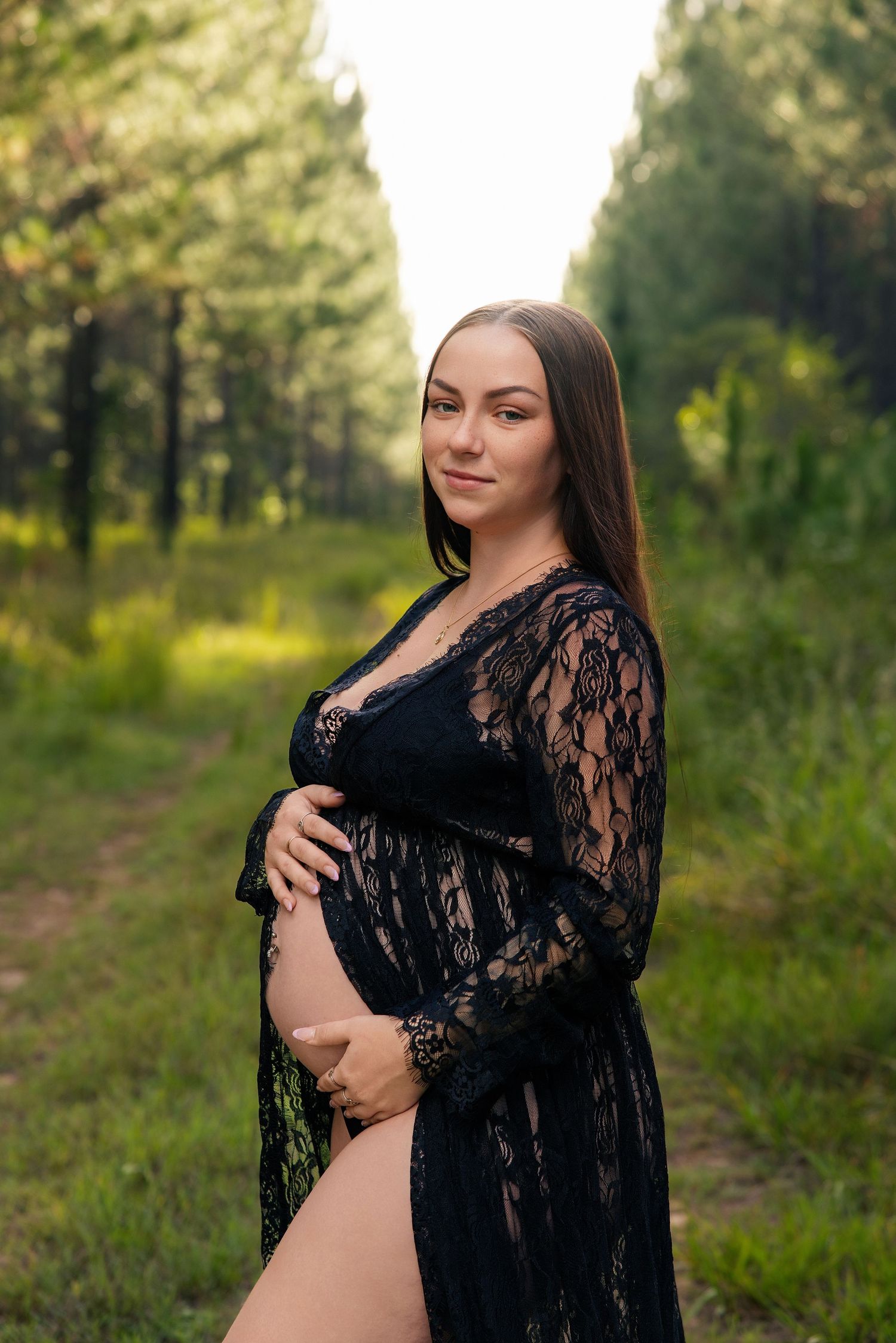 Maternity photo of woman in black lace dress posing outdoors in a pine forest setting.