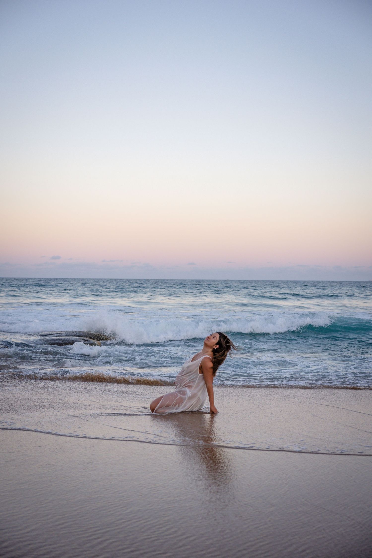 A graceful figure in a flowing dress poses on a beach at sunset as ocean waves crash on the shore.